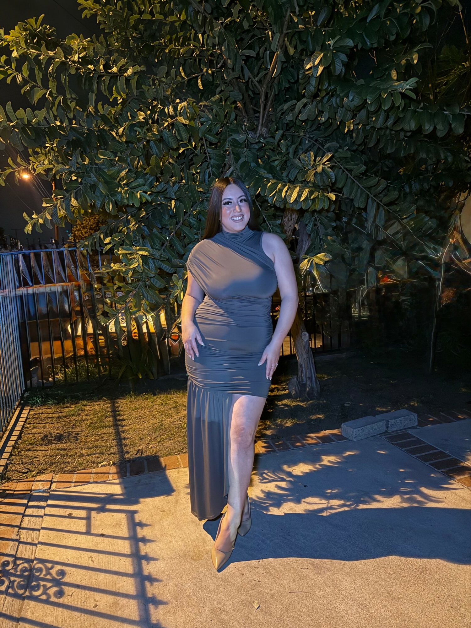 Woman in a long dress standing outdoors at night with trees and a fence in background.