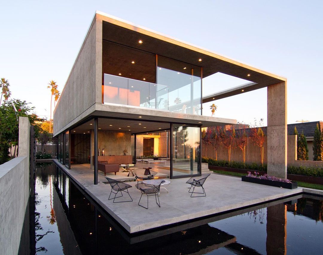 Modern two-story house with large glass windows, outdoor seating area, and reflecting pool, during sunset.