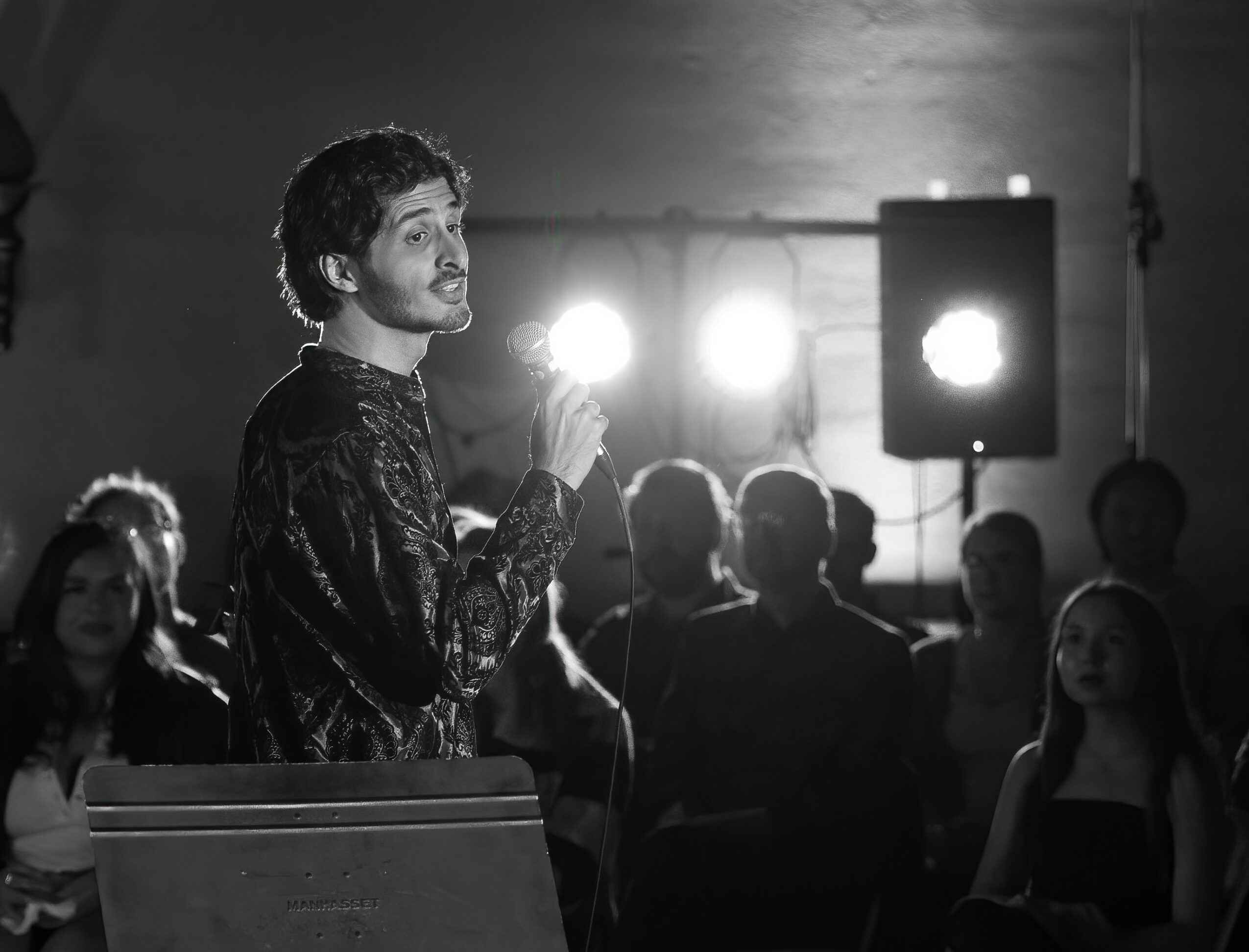 Person holding microphone speaking to audience in a dimly lit room with bright stage lights behind.
