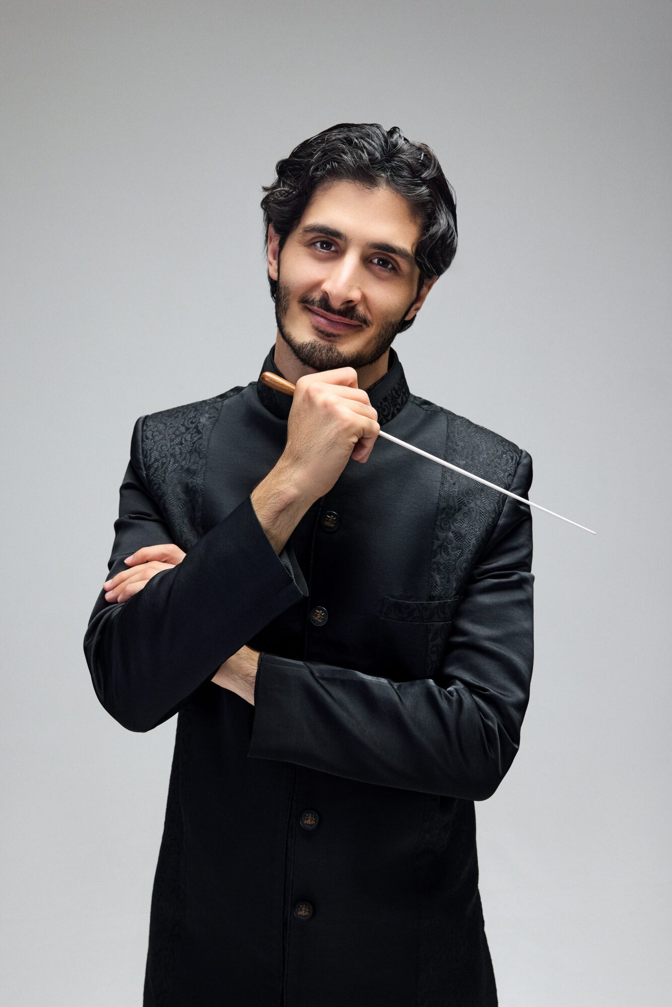 Young man with dark hair and beard, wearing a black traditional outfit, holding a conductor's baton, smiling.
