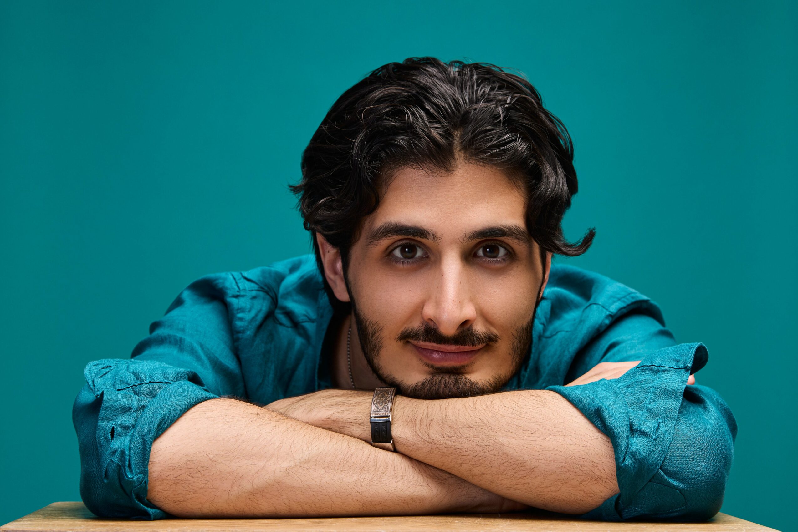 Man with dark hair and beard resting his arms on a table, wearing a blue shirt, against a teal background.