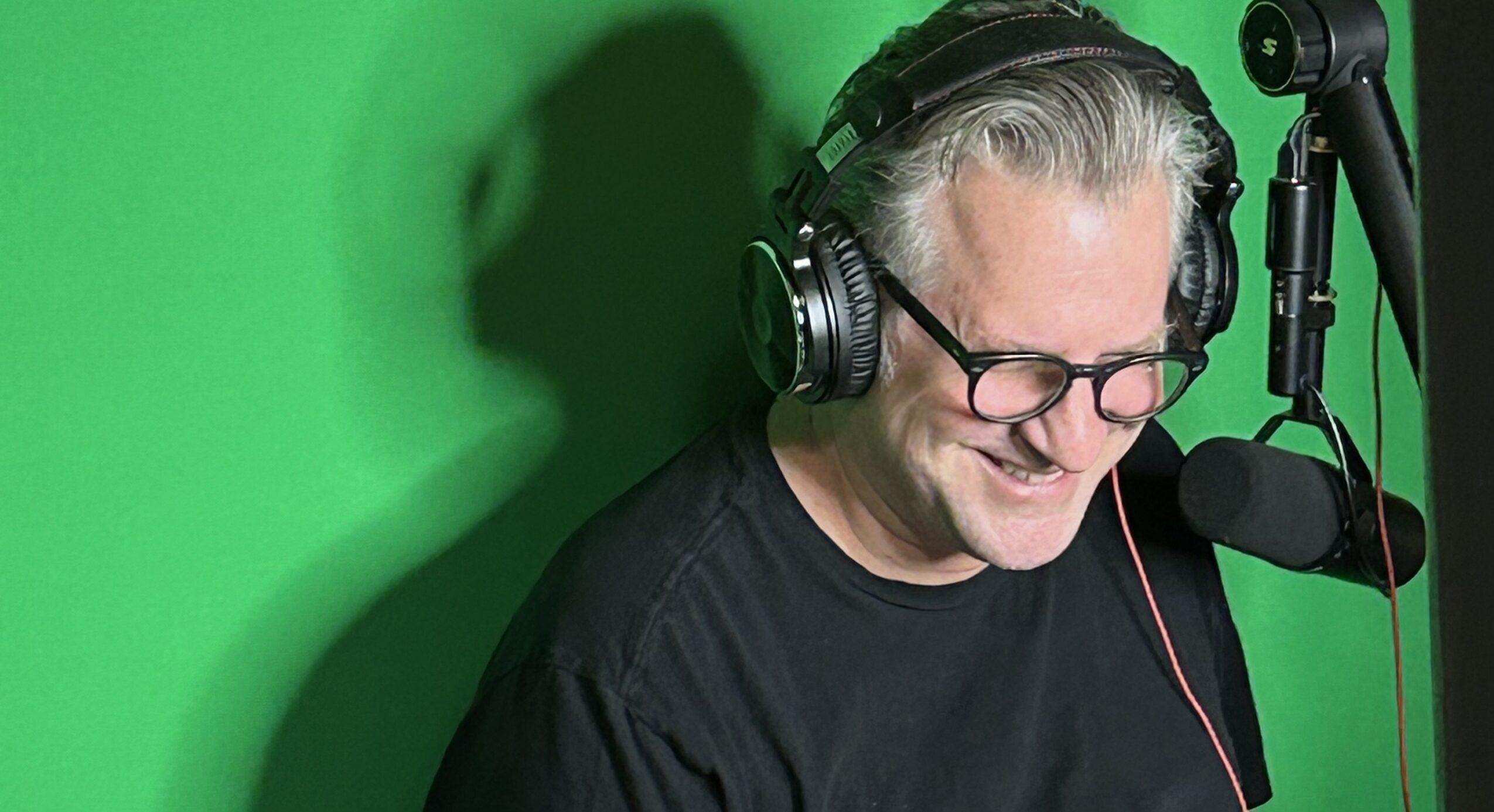 Older man with glasses and headphones smiling in front of a green screen, speaking into a microphone.
