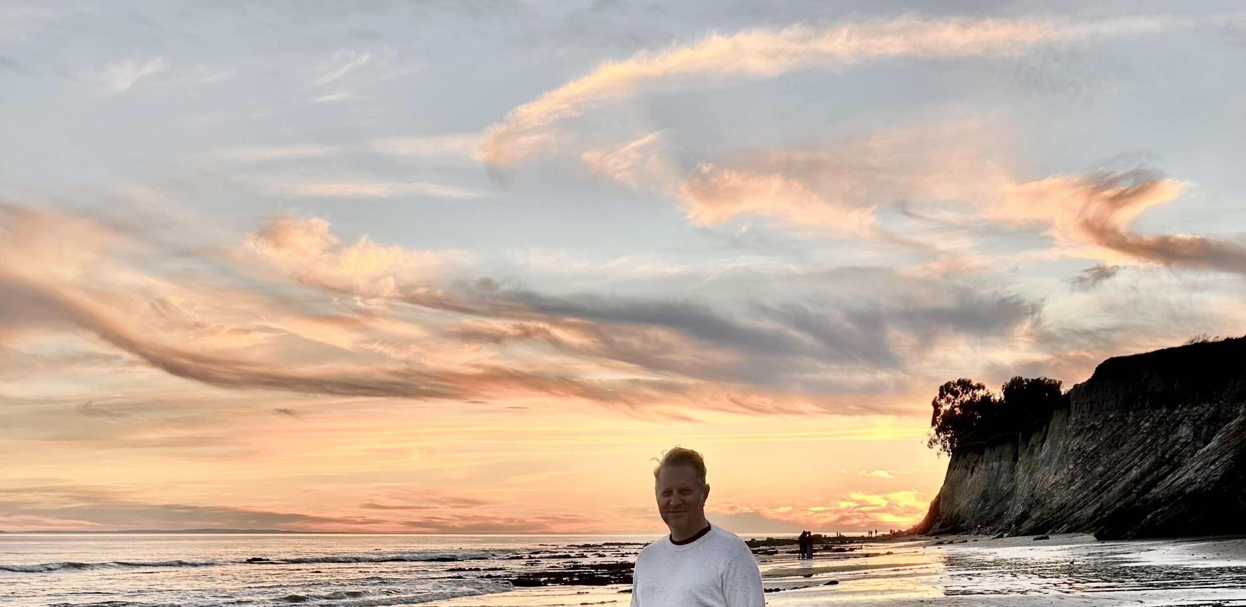 Person standing on beach near water with cliffs and colorful sky at sunset.
