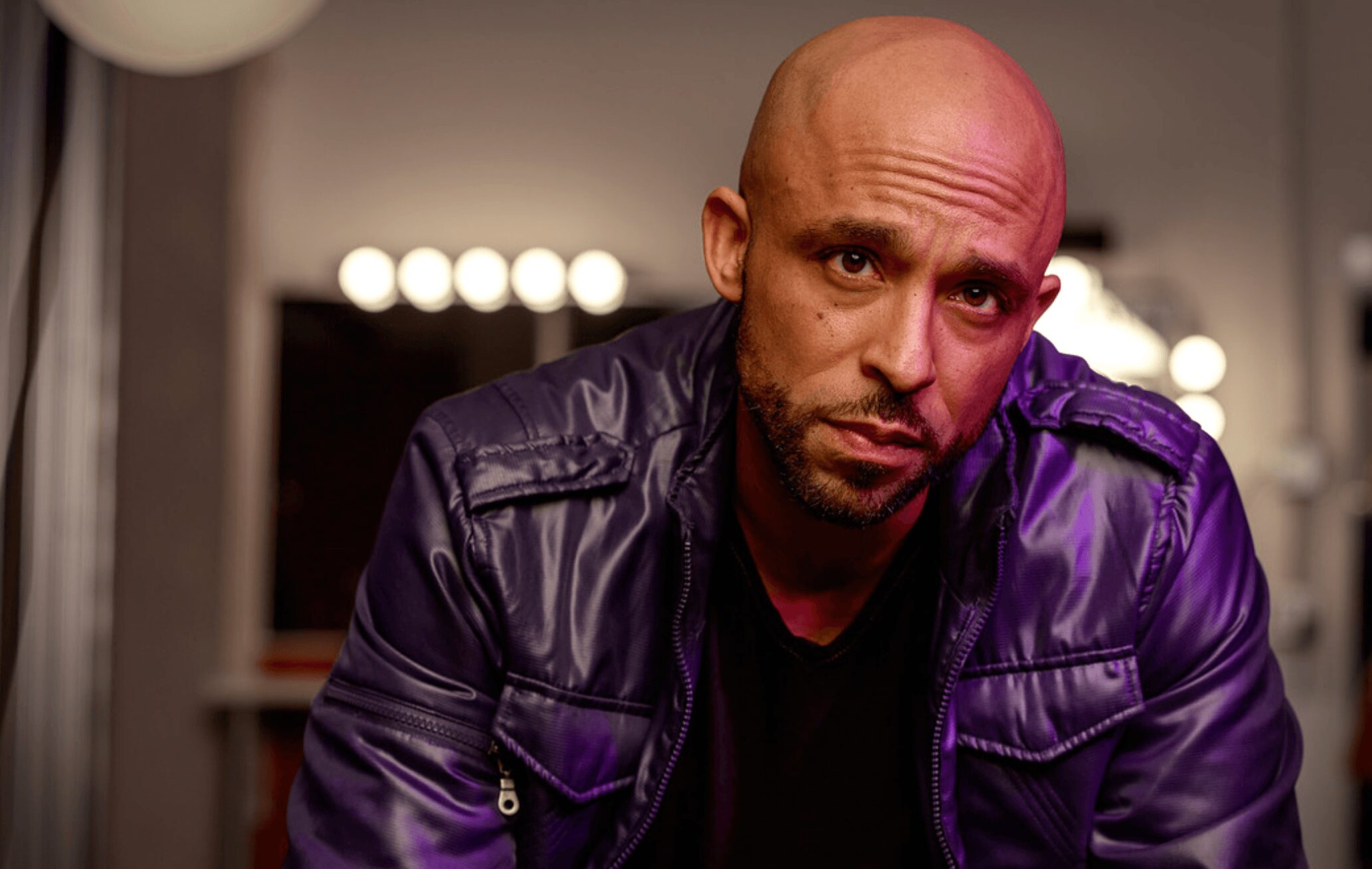 Man with a bald head and beard wearing a dark jacket, looking at the camera indoors.