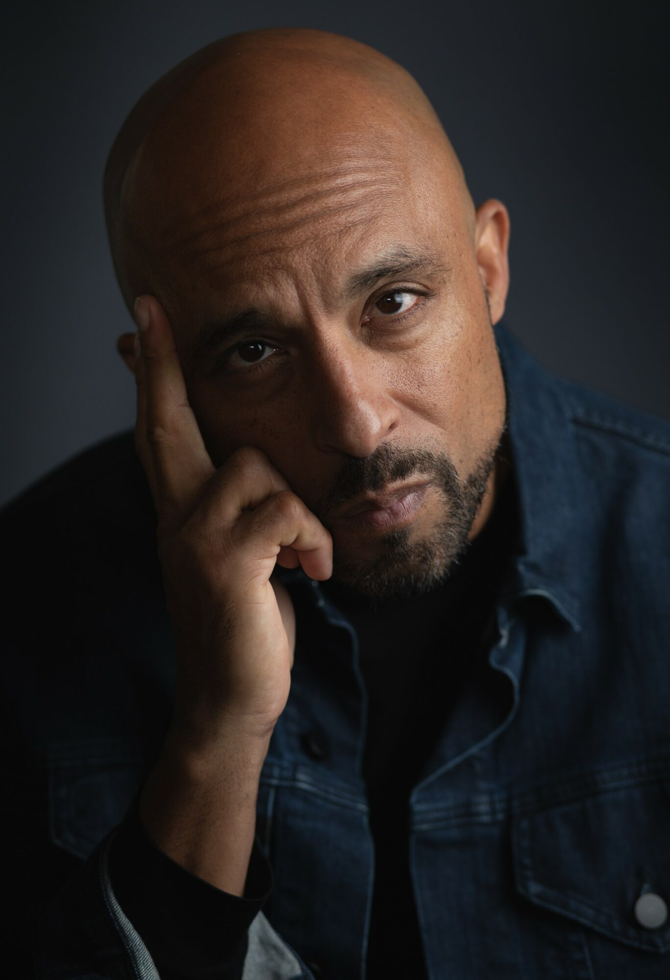 Bald man with a beard resting his head on his hand, wearing a dark shirt, against a dark background.
