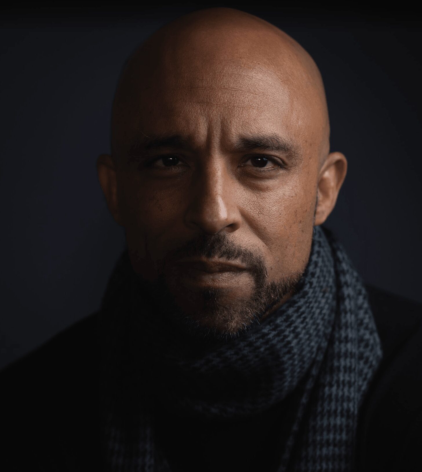 Close-up portrait of a bald man with a serious expression, wearing a dark scarf against a dark background.