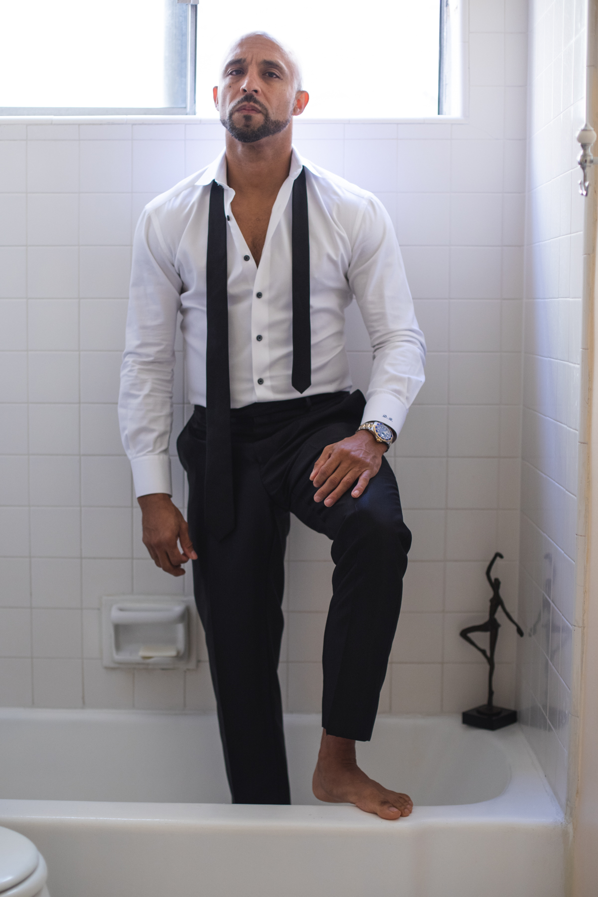 Man standing in a bathroom with one foot on the bathtub edge, wearing formal attire, looking up.