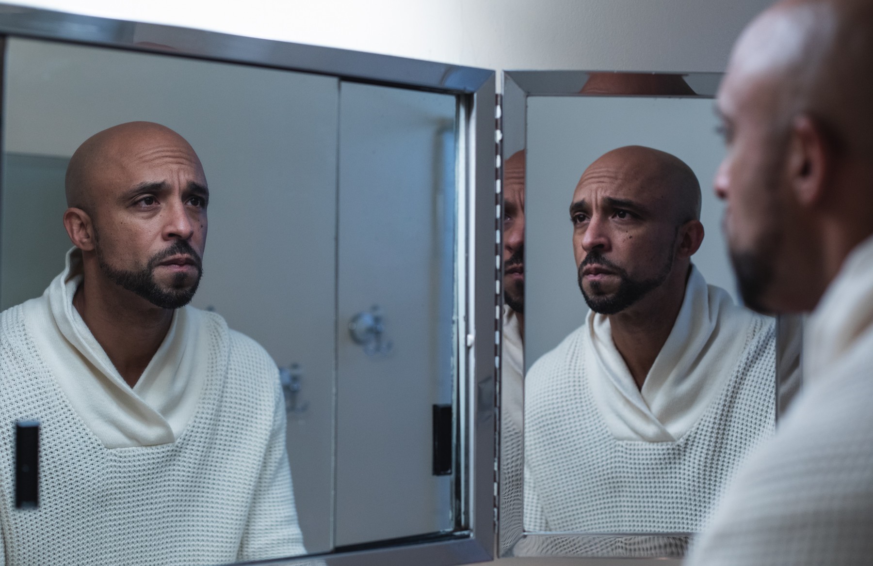 Man with a shaved head and beard looking at his reflection in a mirror, wearing a white robe.
