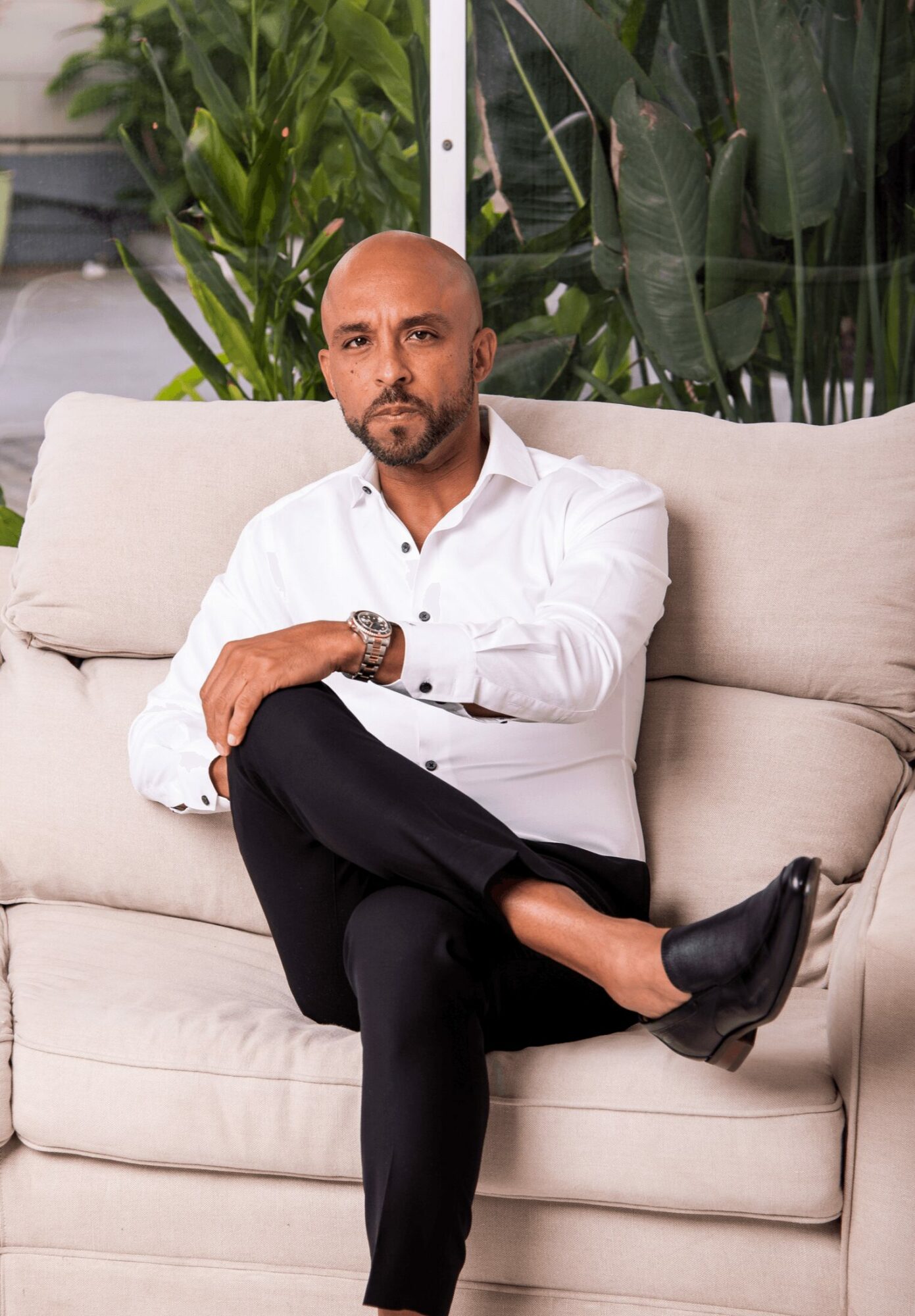Man with a beard and bald head sitting on a beige sofa, wearing a white shirt and black pants, with plants behind him.