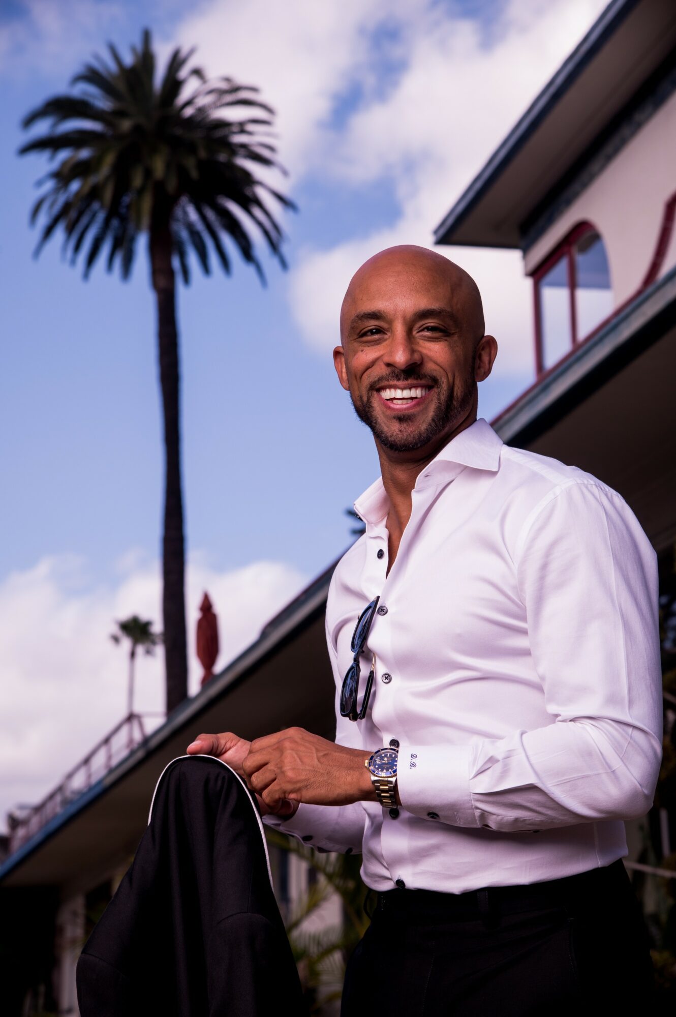 Smiling man in white shirt outdoors with palm tree and building in background.