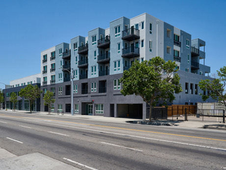 Modern multi-story apartment building with balconies, trees, and a street in front, under a clear blue sky.