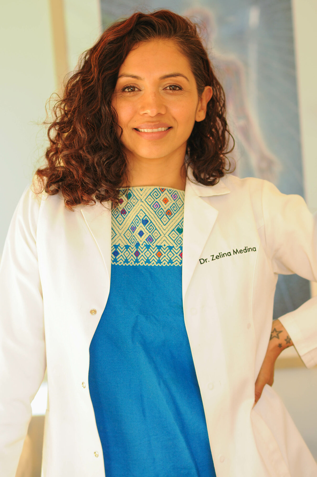 Young woman with curly hair wearing a white coat and colorful top, smiling indoors.