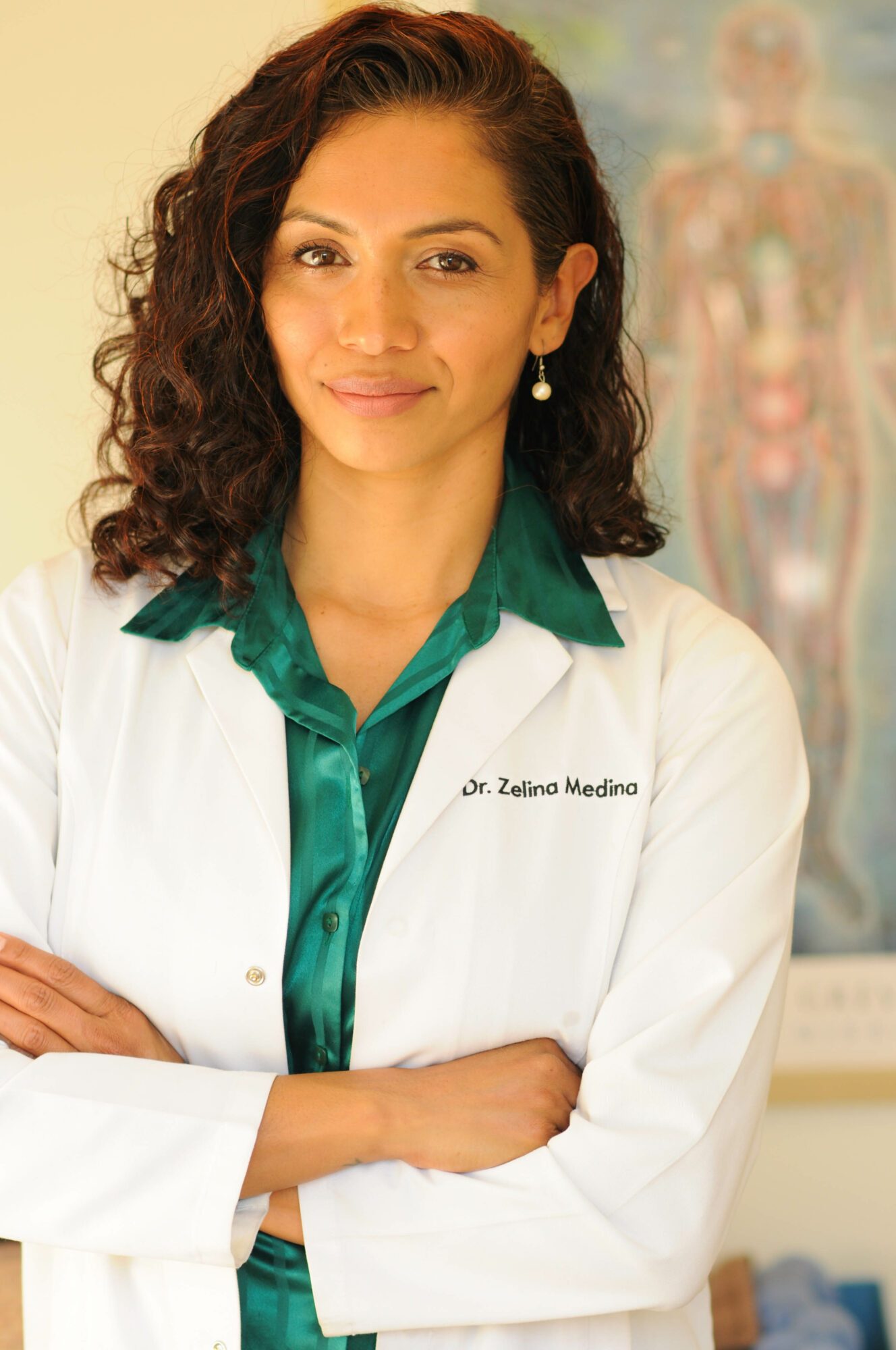 Woman with curly hair in white coat and green shirt, standing in medical office, smiling.
