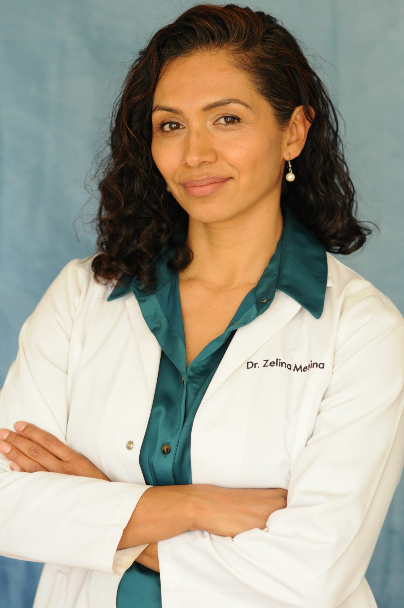 Woman with dark curly hair wearing a white lab coat and teal shirt, smiling with arms crossed.