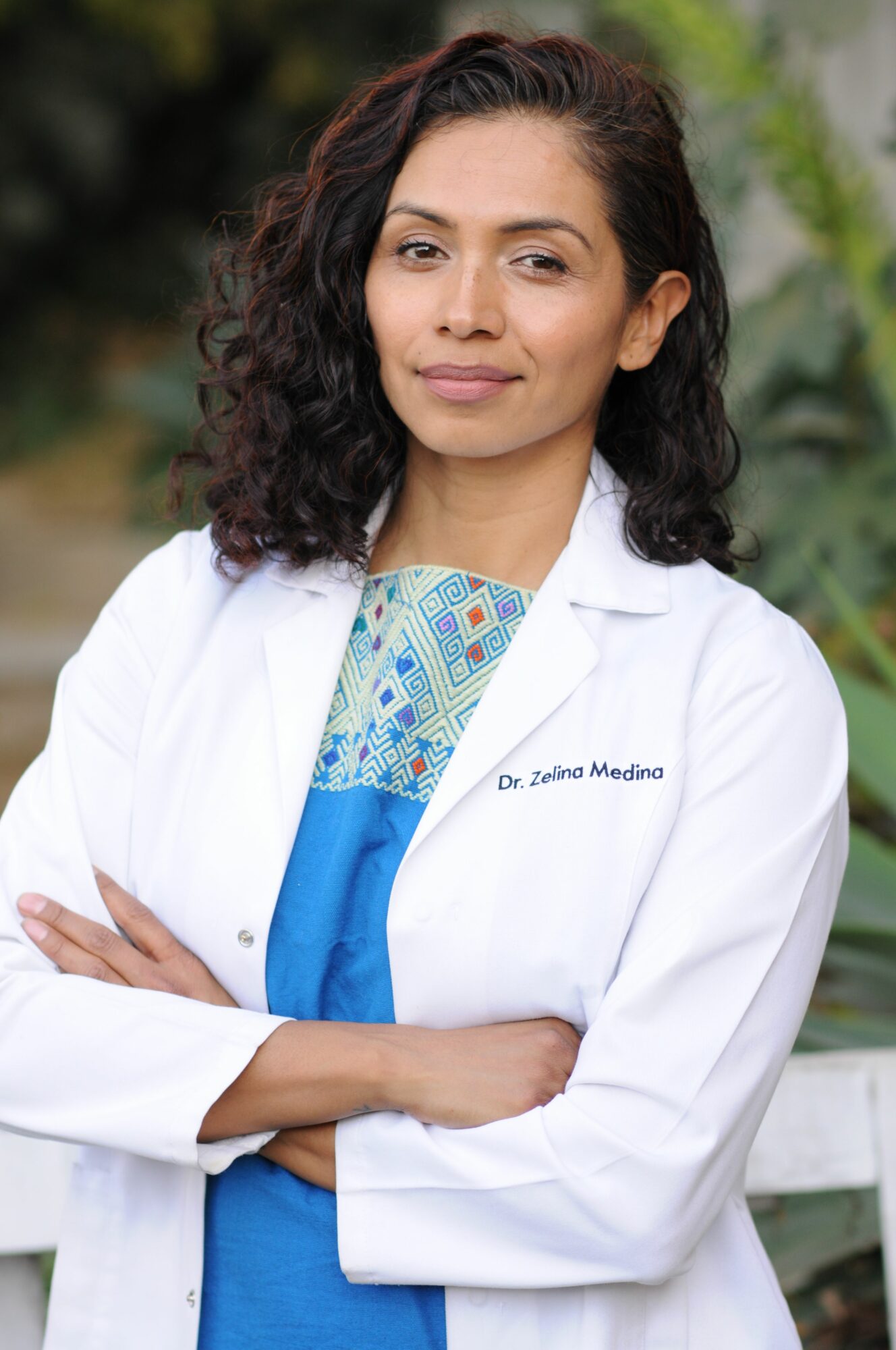 Woman in white coat with arms crossed, outdoors, smiling, wearing patterned top and blue dress.