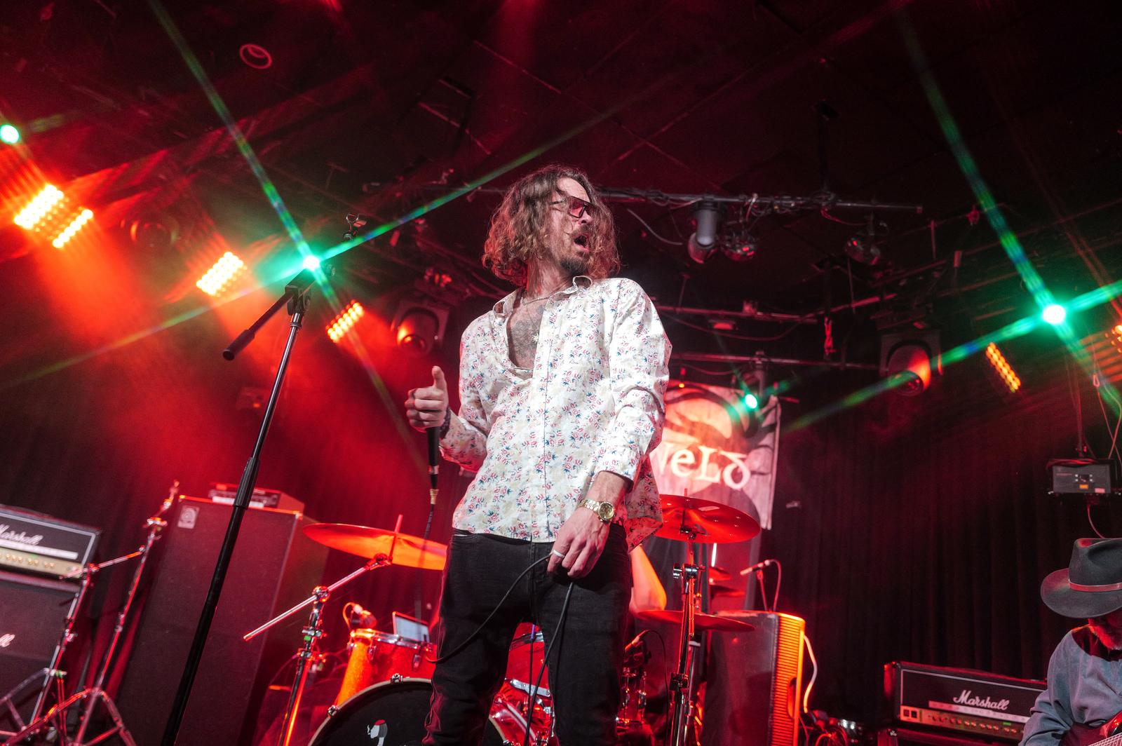 Person with curly hair holding a microphone on stage with colorful lights and musical equipment.