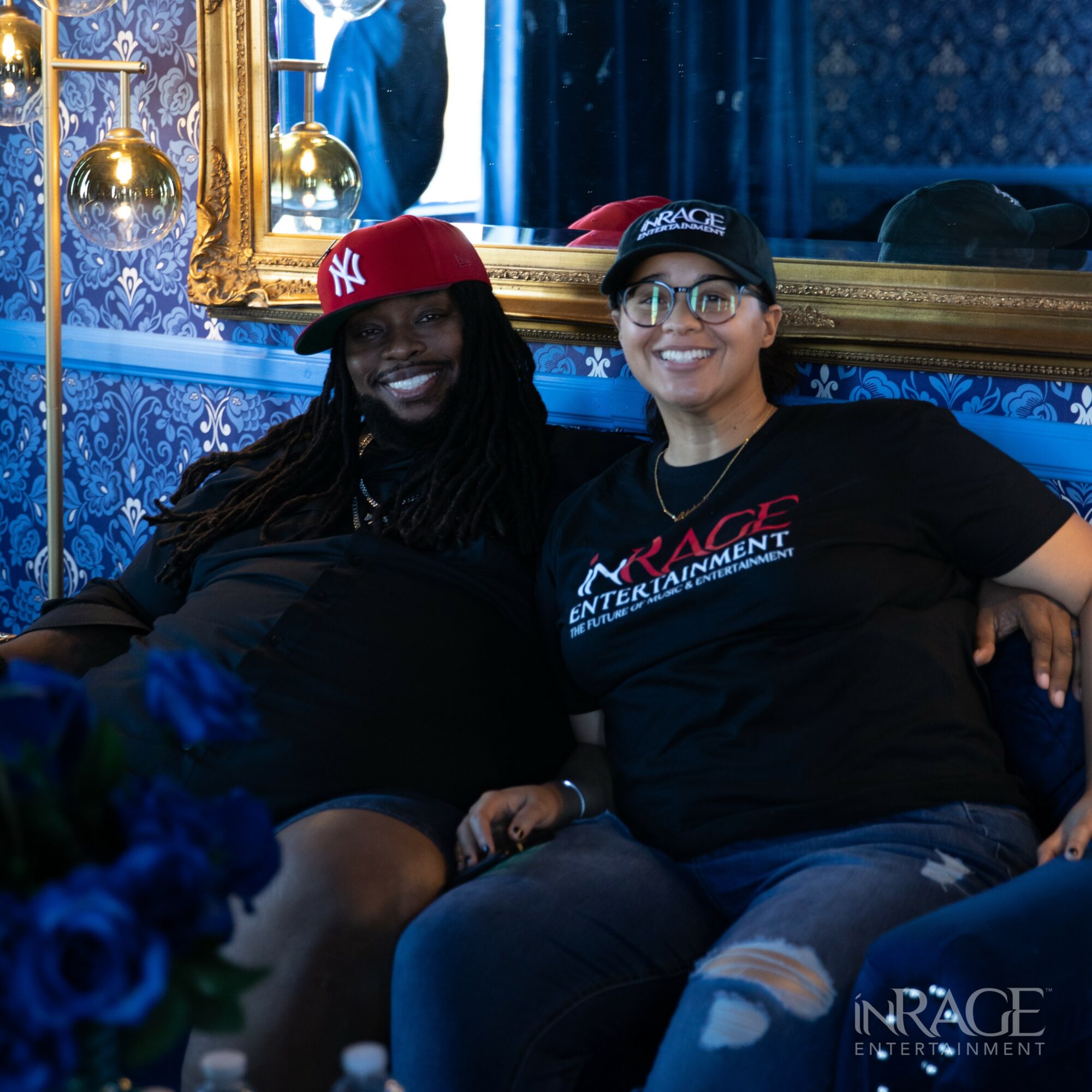Two women sitting on a blue couch, smiling, in a room with ornate gold-framed mirror and blue patterned wallpaper.