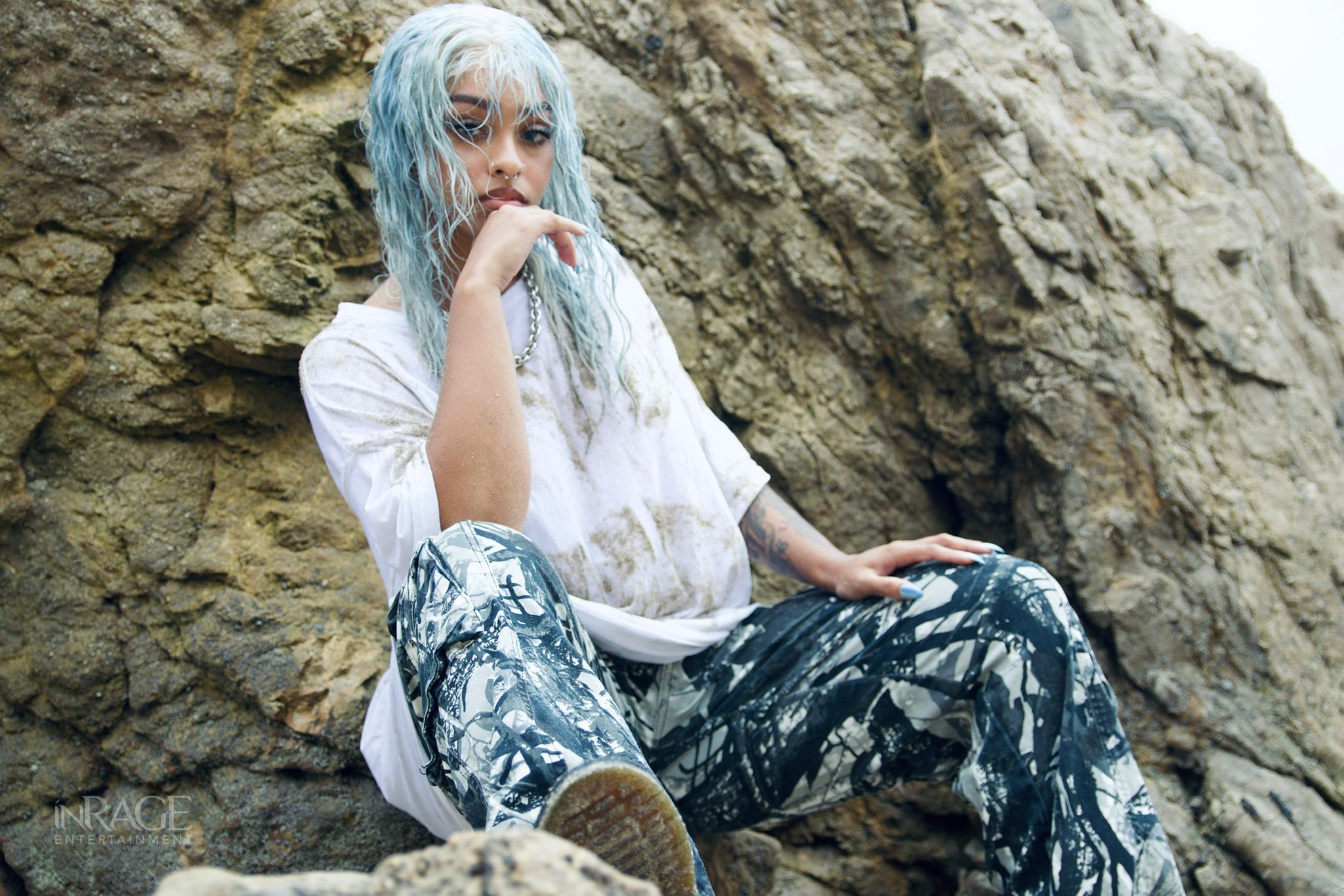 Person with blue hair sitting against a rocky background, wearing a white shirt and patterned pants, with hand near face.