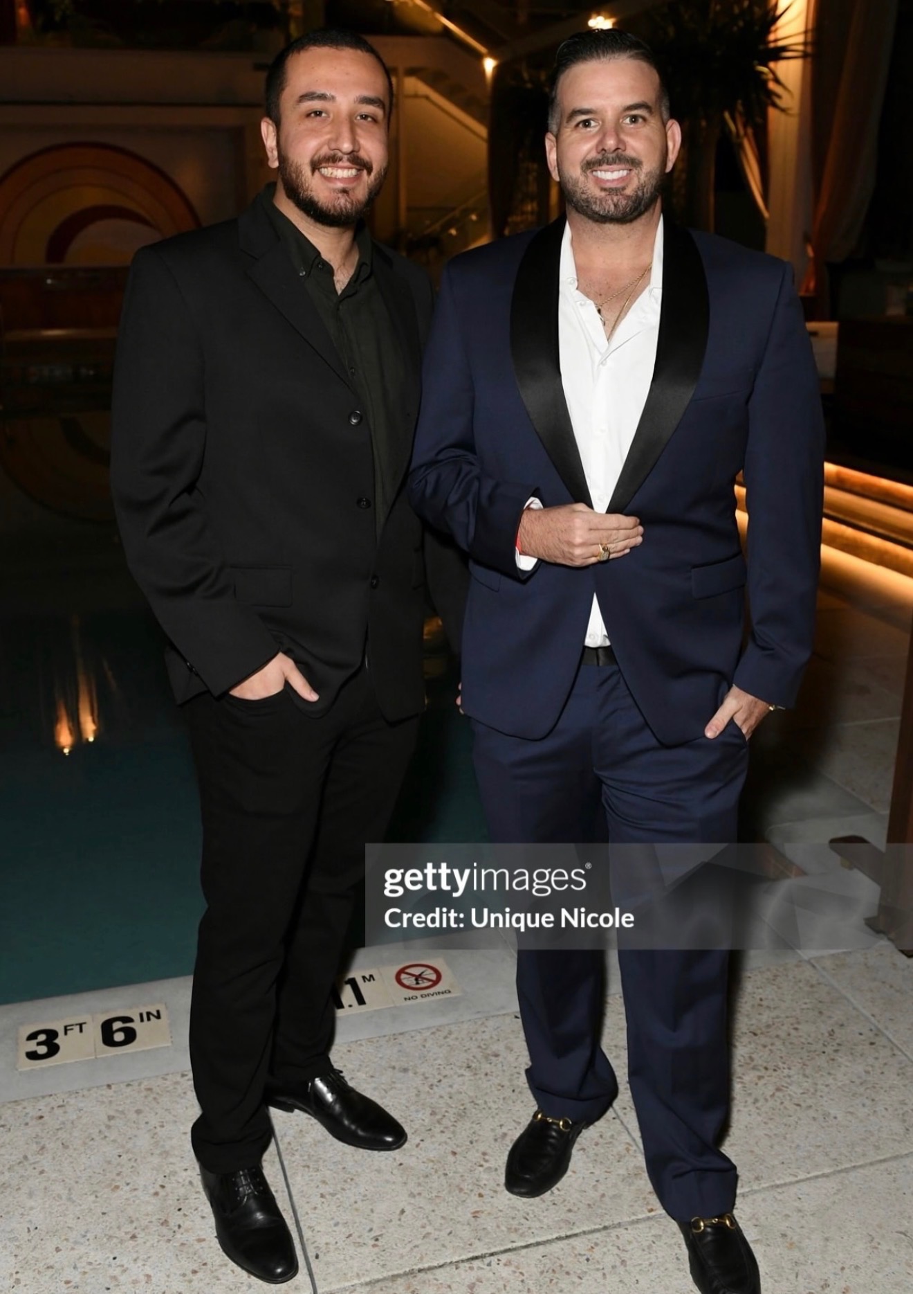 Two men in suits standing indoors, smiling, with warm lighting and decorative background.