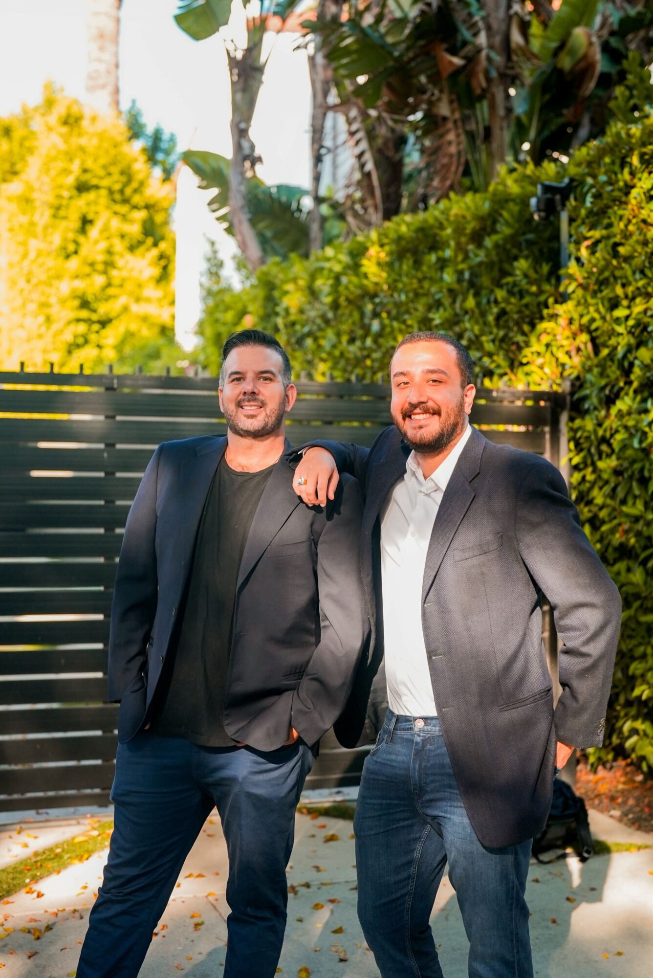 Two men in suits standing outdoors, smiling, with greenery and a fence in the background.