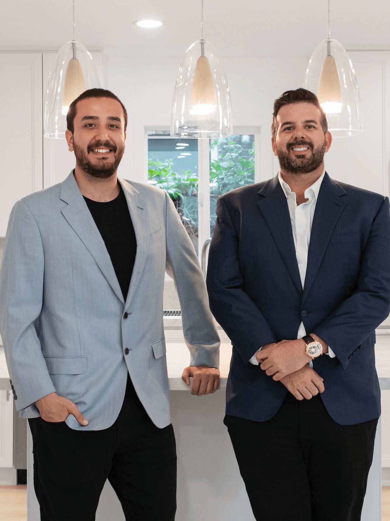 Two men in suits standing indoors with hanging lights and a window behind them.