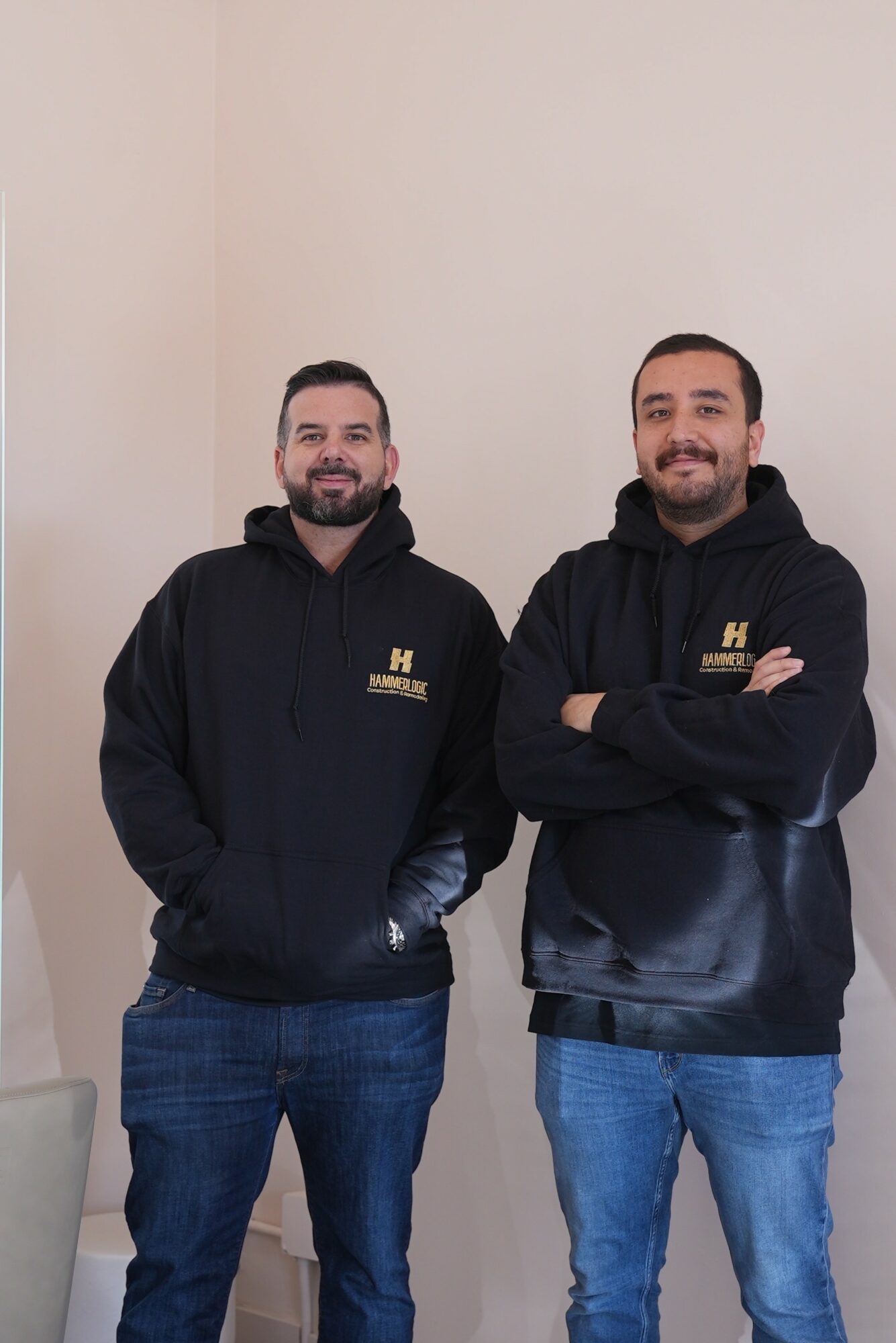 Two men standing side by side, both wearing black hoodies with a logo, smiling, with arms crossed or hands in pockets.