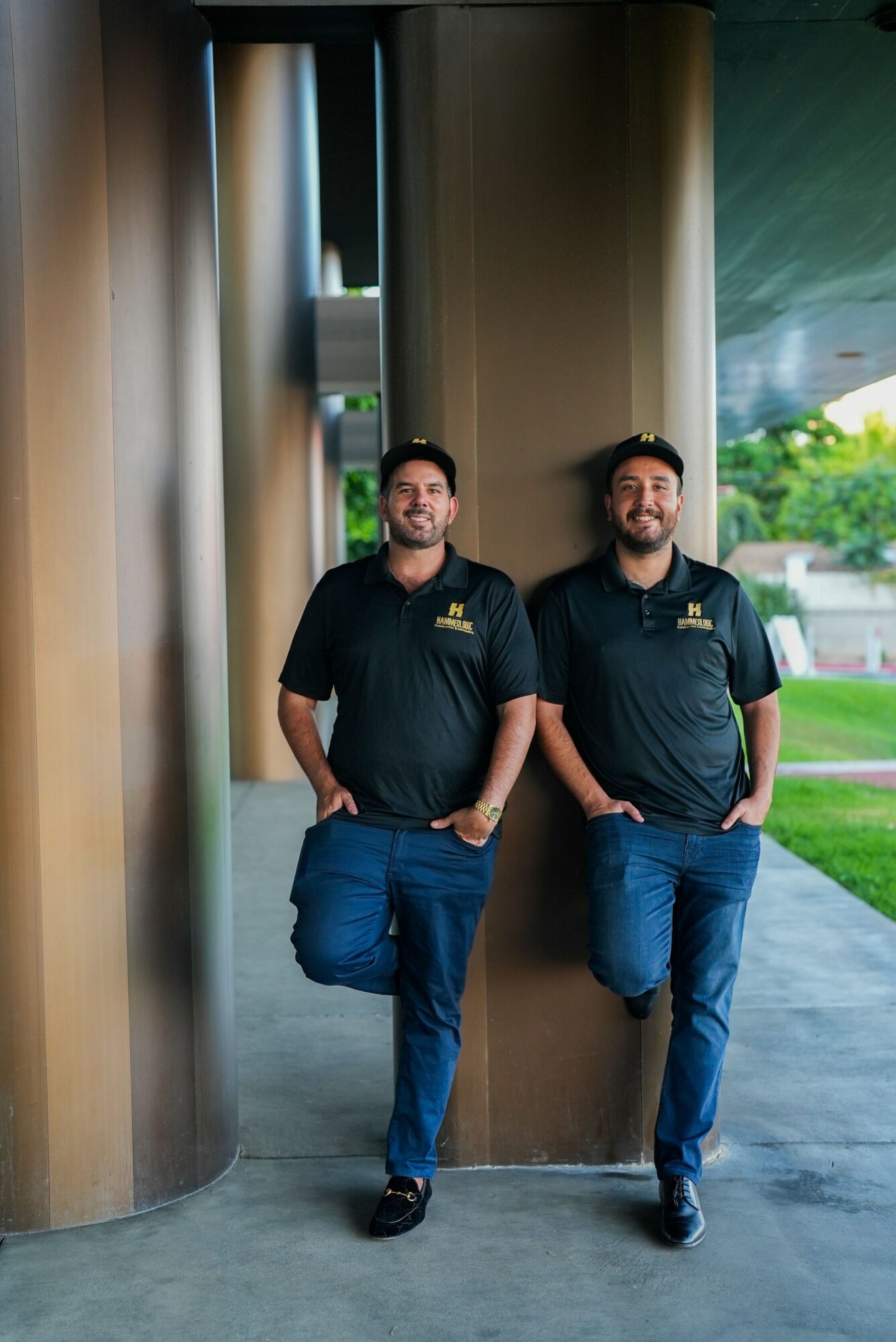 Two men in black shirts and jeans leaning against a metallic wall outside, smiling, with greenery in the background.