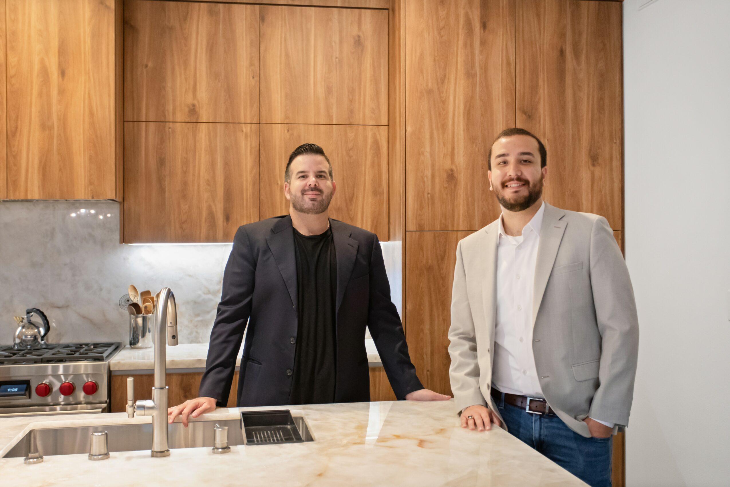 Two men standing behind a kitchen counter with wooden cabinets in the background.