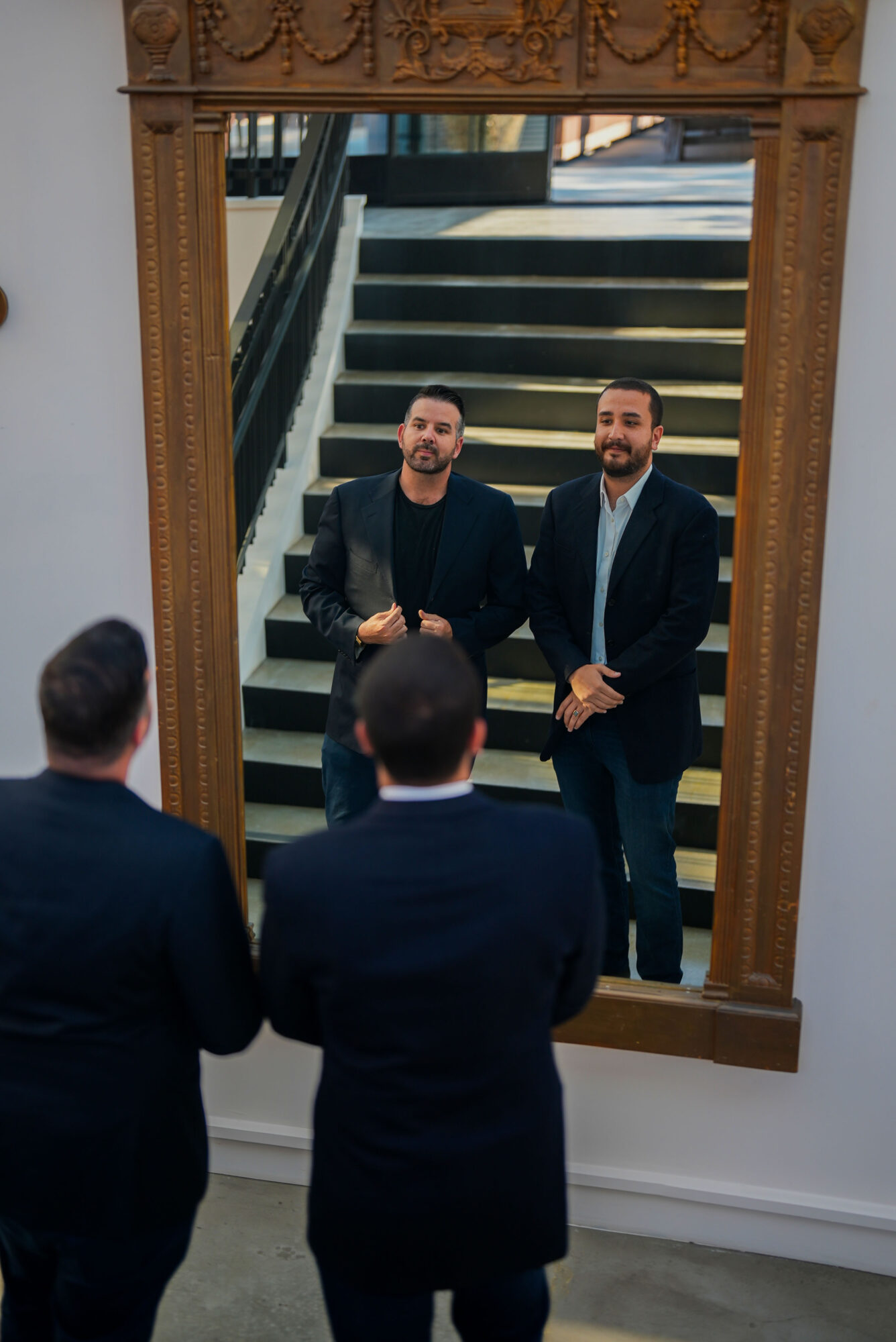 Two men in suits stand in front of a mirror, looking at their reflection, with three people facing them in the foreground.