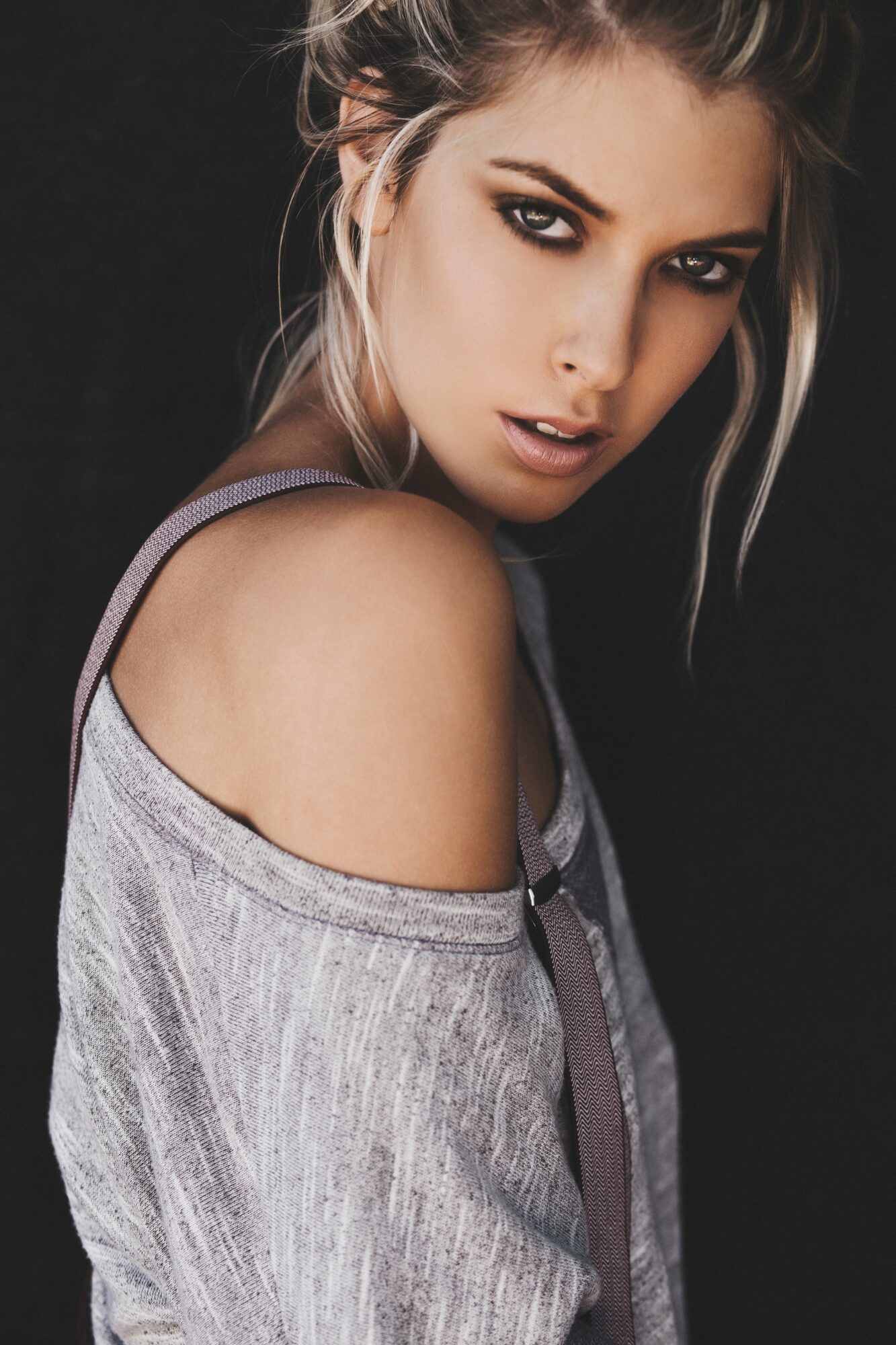 Young woman with makeup and loose hair, wearing a gray off-shoulder top, looking at camera against black background.