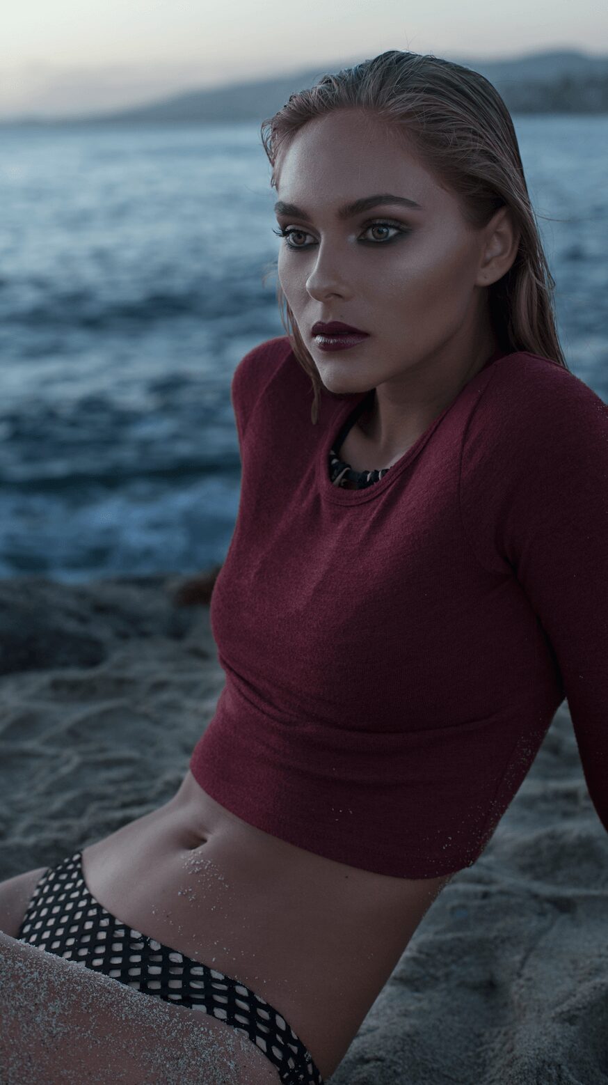Young woman with makeup and long hair, sitting on a beach near water, wearing a maroon top and polka dot shorts.