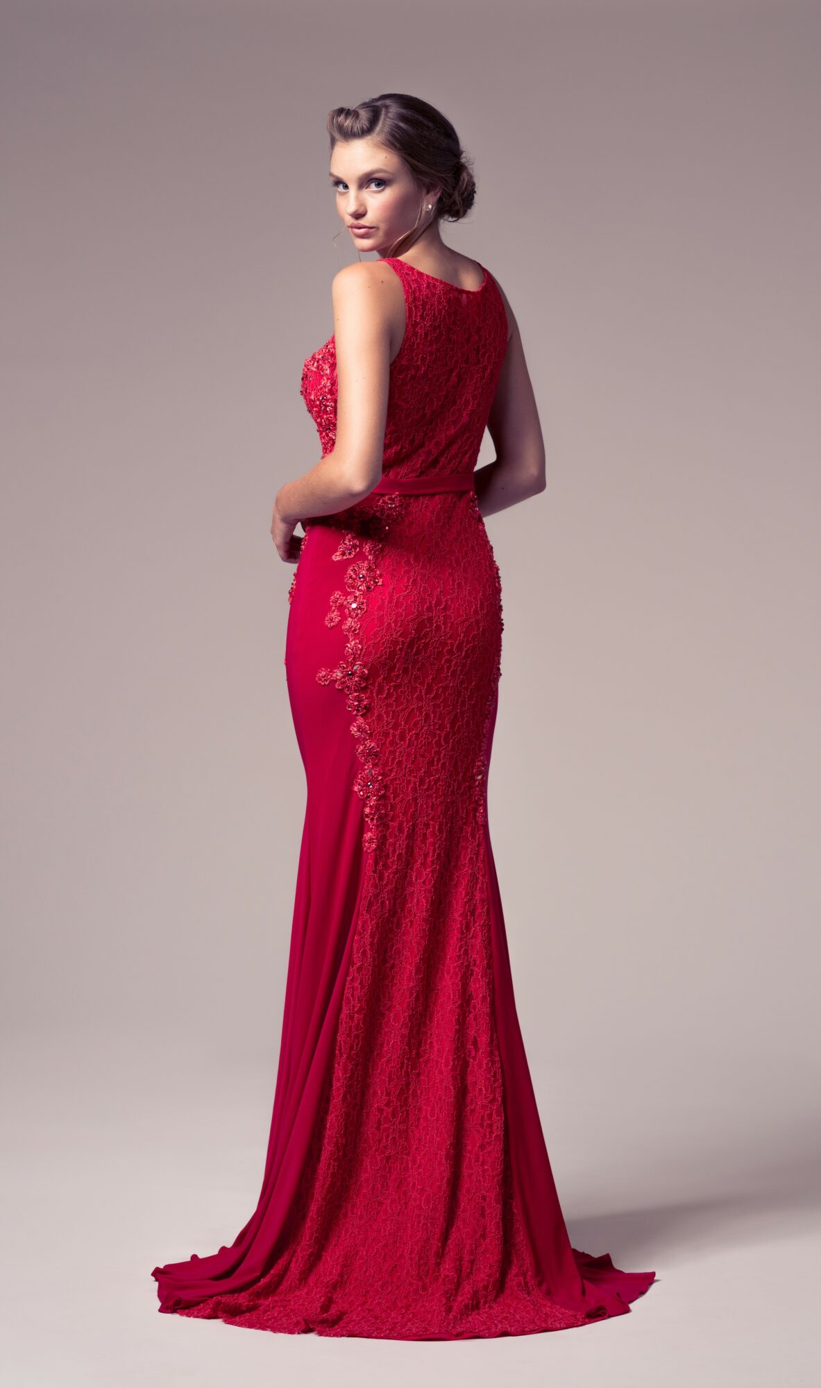 Woman in a long red lace evening gown, standing sideways with her head turned towards the camera.