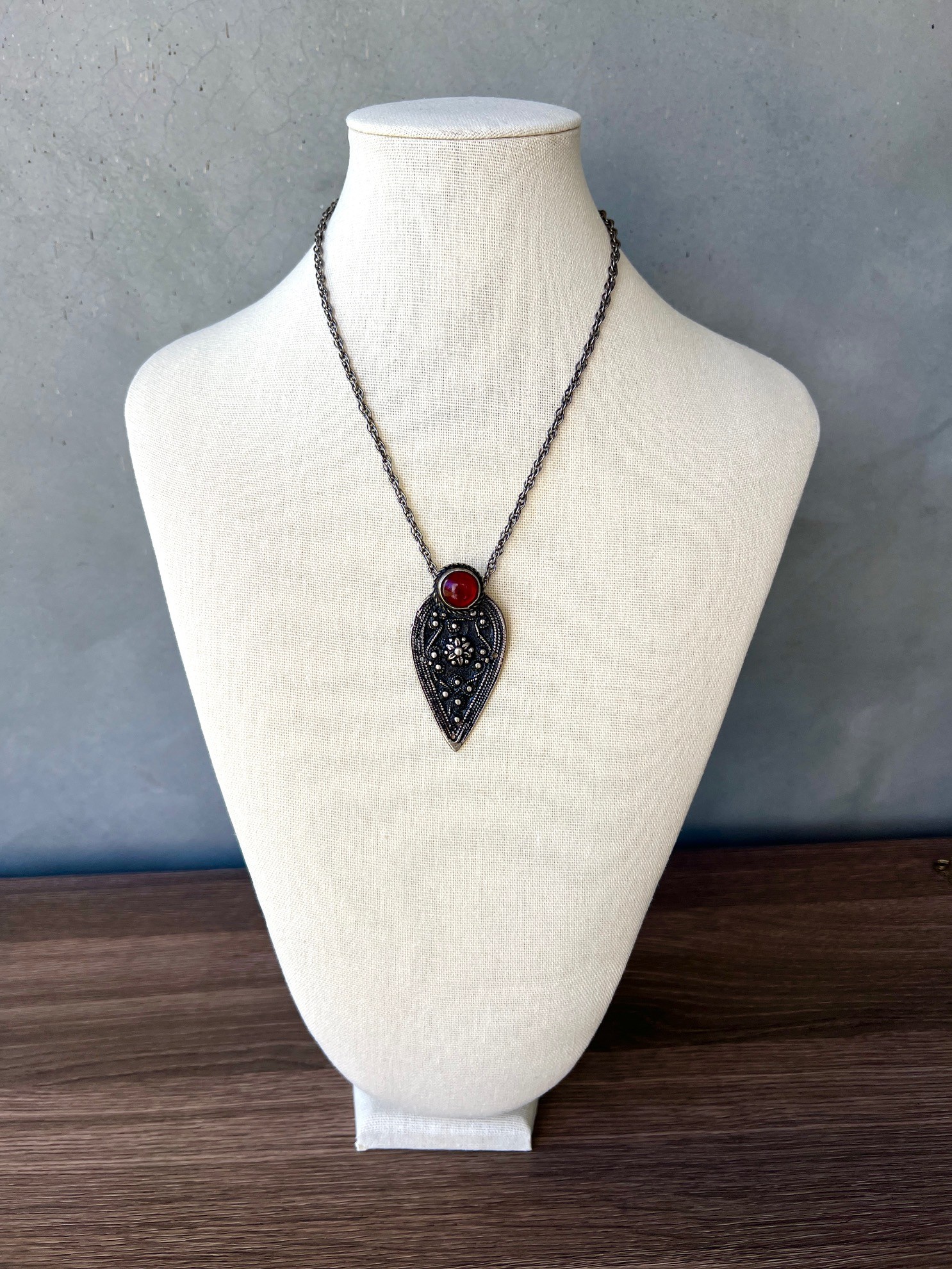 Pendant necklace with a red gemstone on a chain displayed on a white bust.