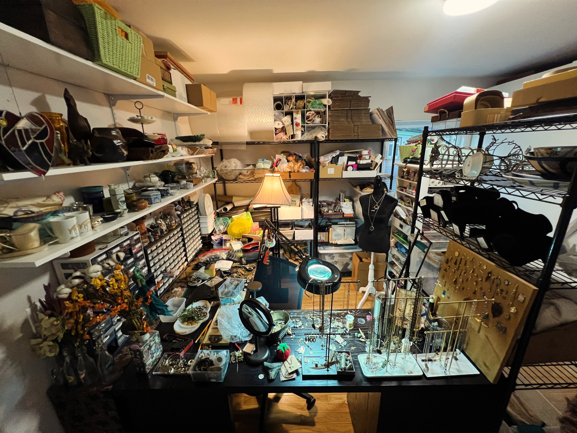 Room filled with shelves and tables holding various household items, including dishes, lamps, and tools, in a cluttered space.