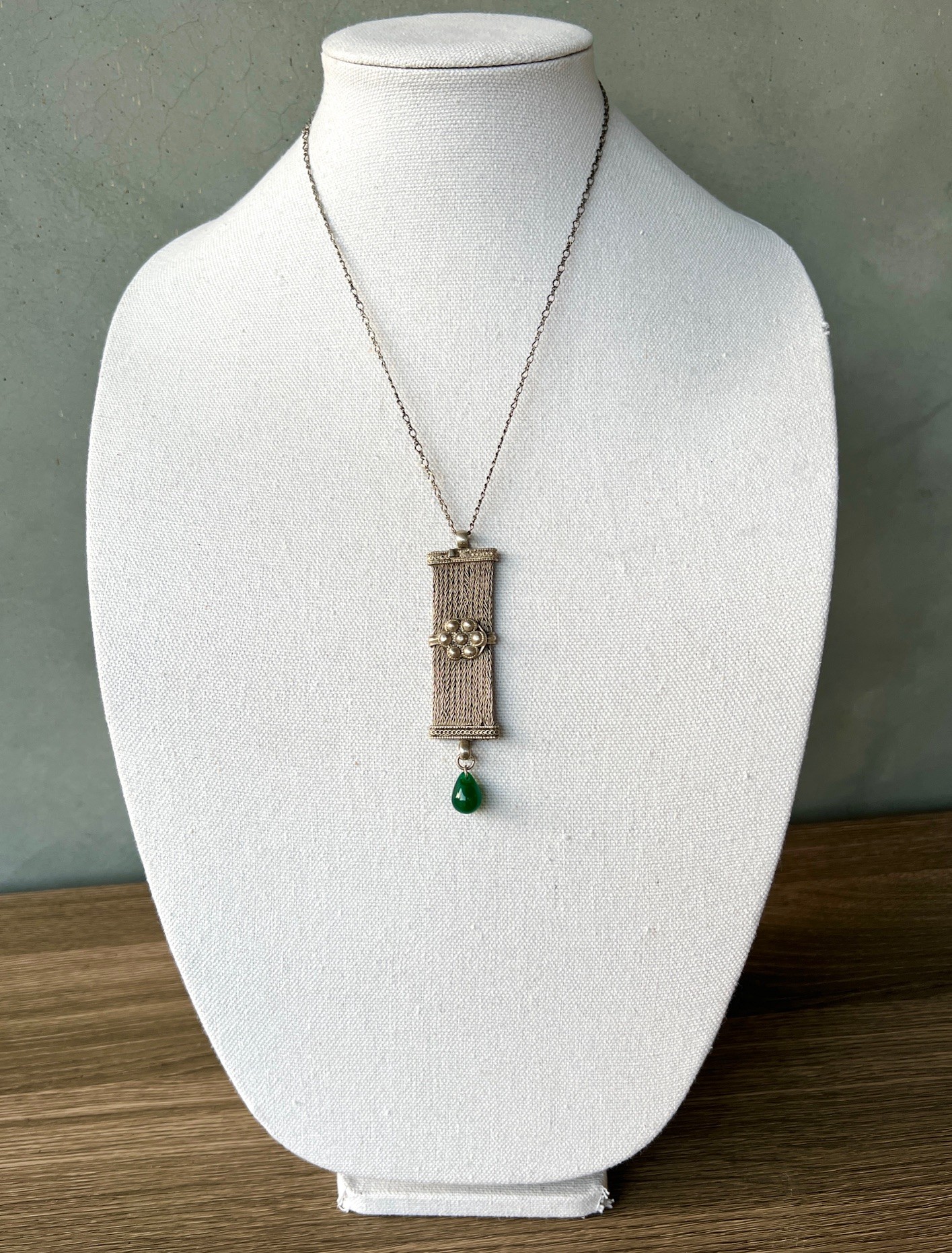 Necklace with a rectangular pendant and a green bead hanging below, displayed on a white bust.