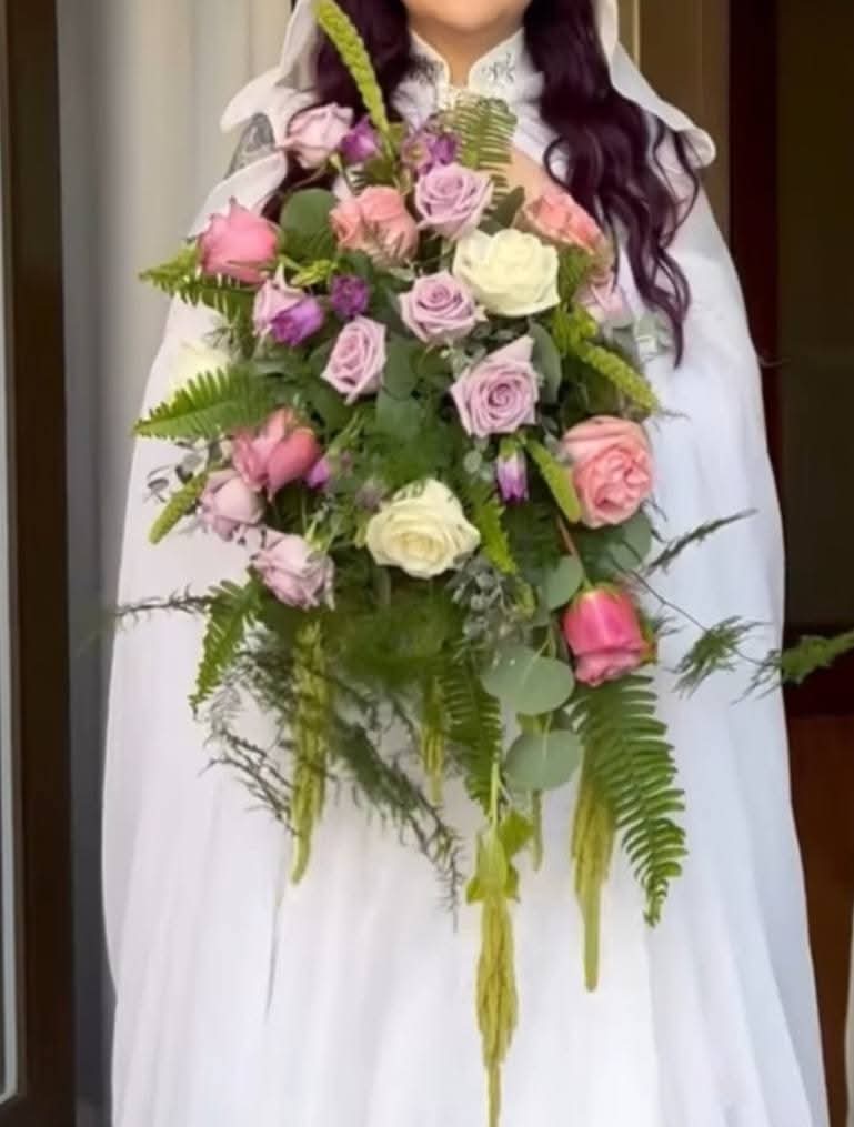 Person holding a large bouquet of pink, purple, and white roses with green foliage, wearing a white dress.