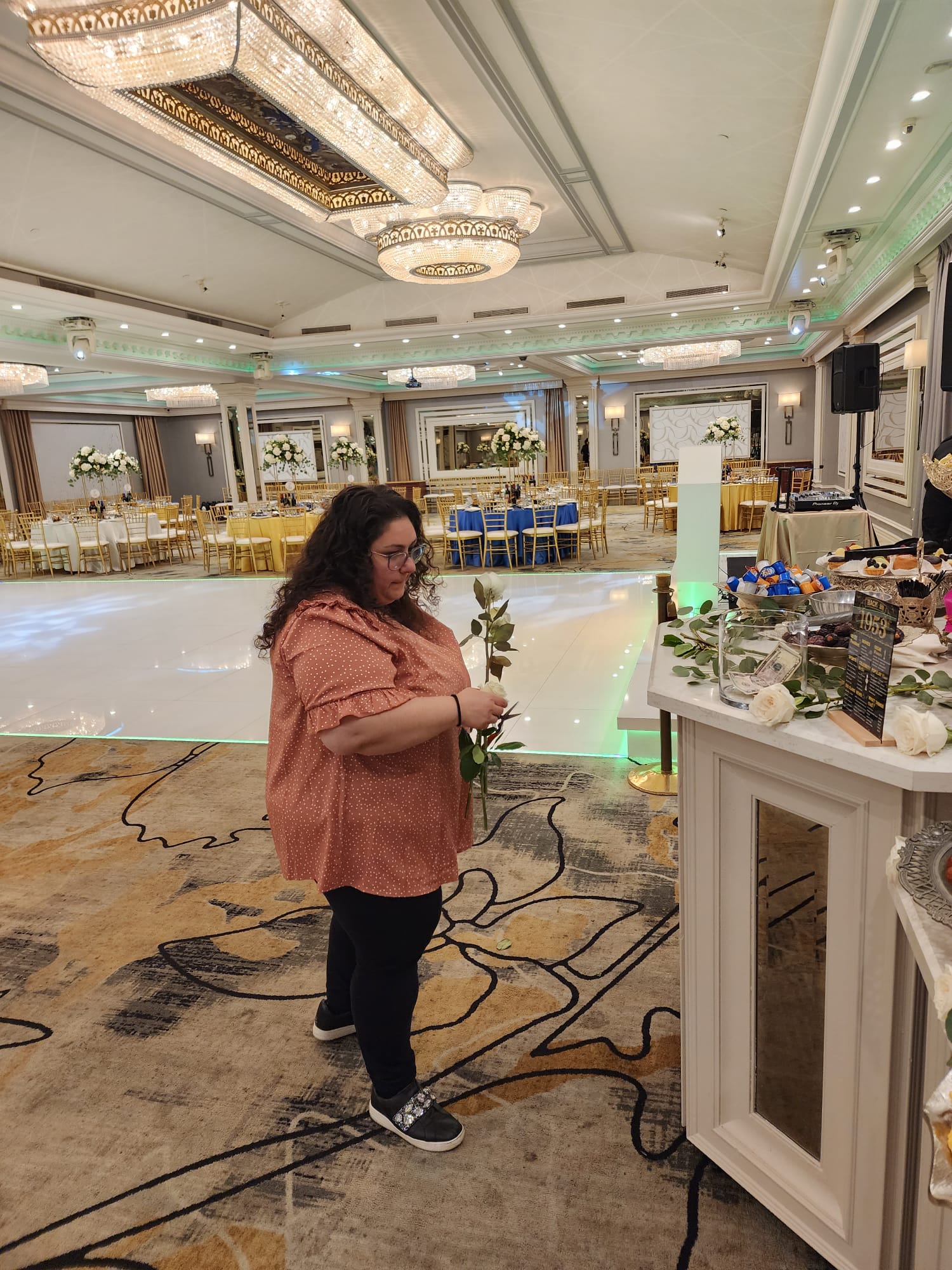 Woman in pink jacket holding a flower in a large banquet hall with decorated tables and chandeliers.