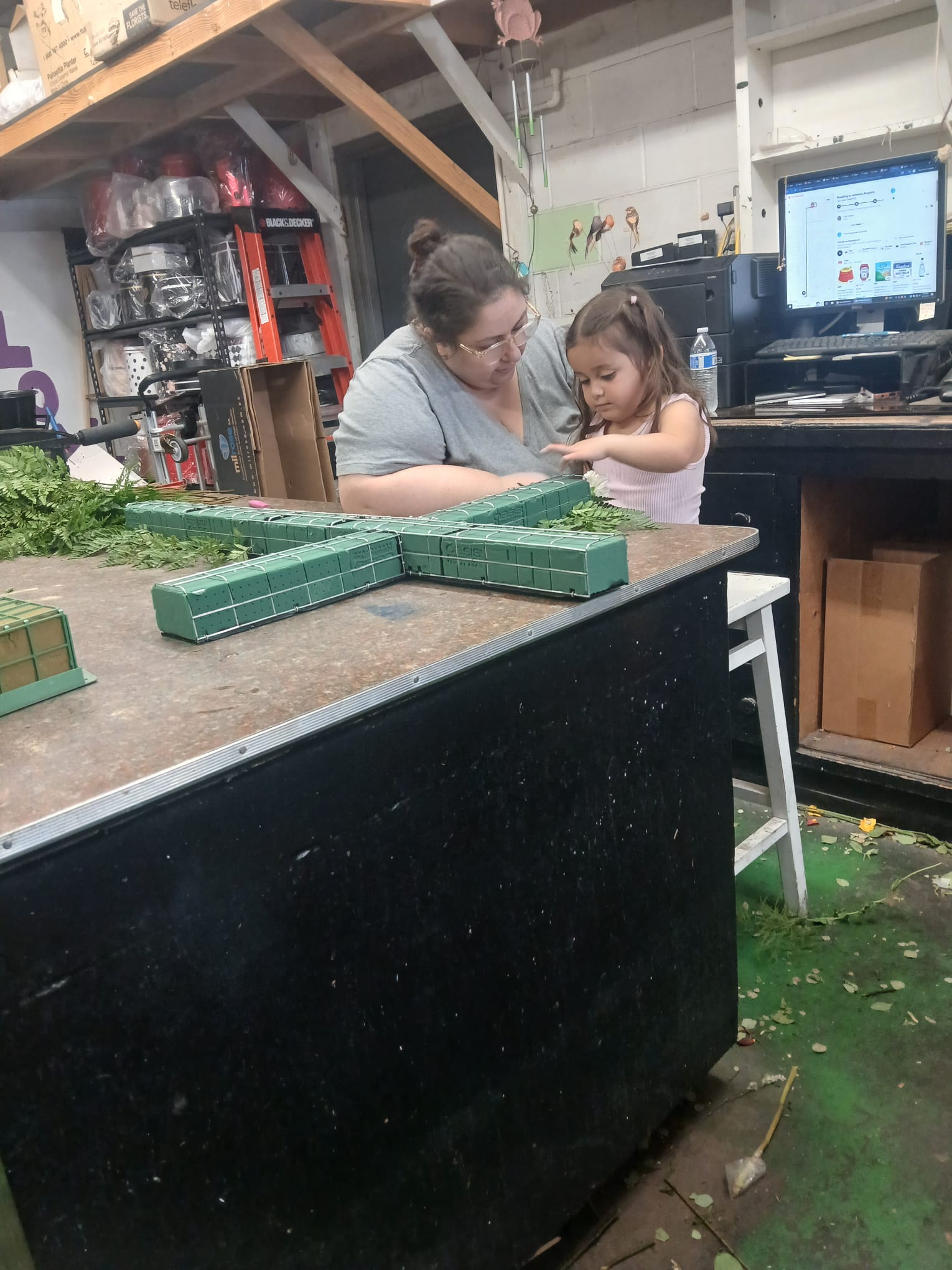 Adult and child working together at a cluttered workbench with green tiles, in a workshop with shelves and a computer.