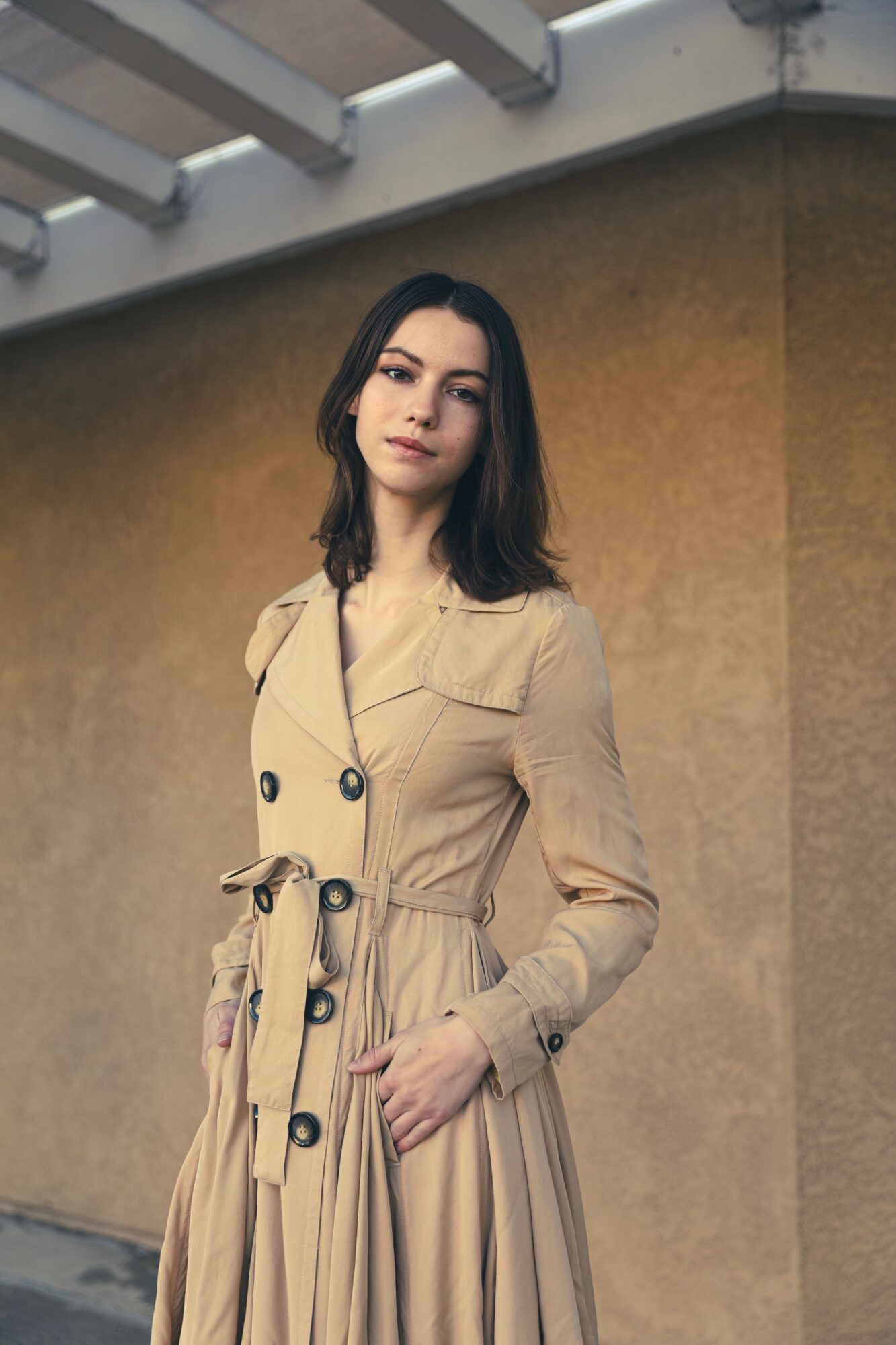 Woman with dark hair in a beige trench coat standing outdoors against a plain wall.
