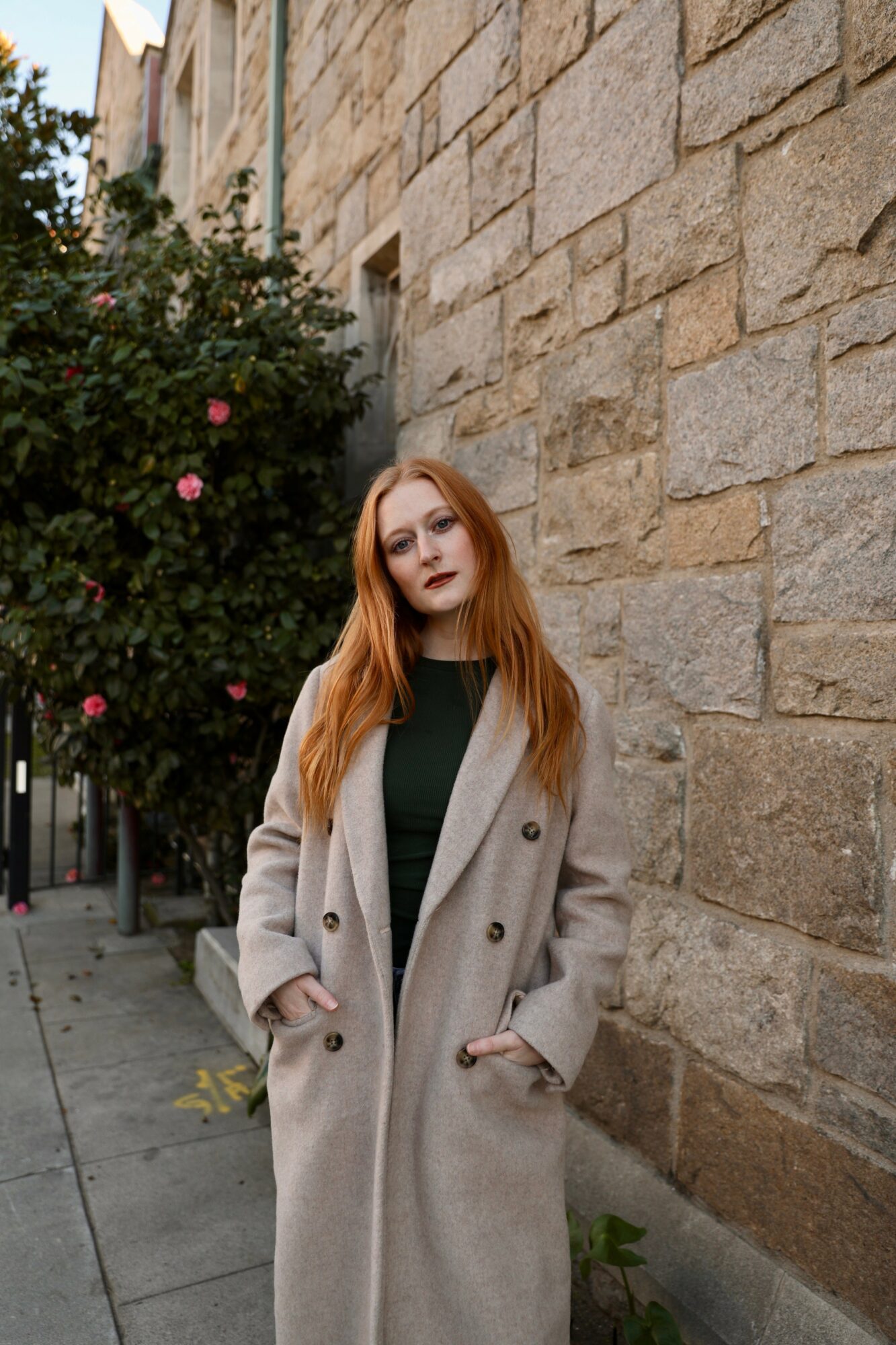 Young woman with long red hair standing against a stone wall and a flowering bush.