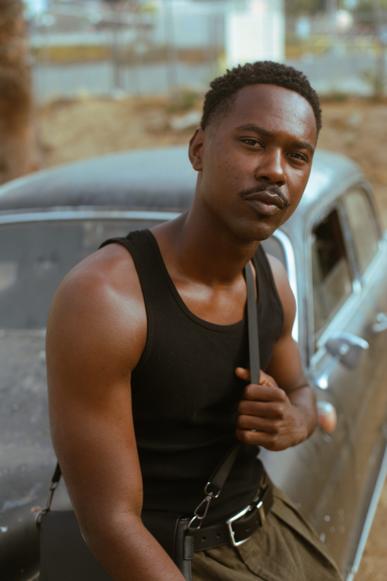 Young man with short curly hair wearing a black sleeveless shirt, standing outdoors near a car.