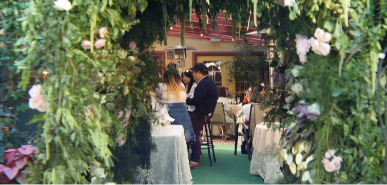 People gathered around tables in a decorated outdoor setting, viewed through a floral archway.