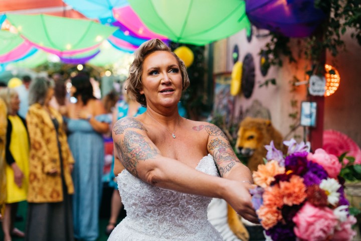 Woman in wedding dress with tattoos on arms, standing outdoors at celebration with colorful umbrellas overhead.
