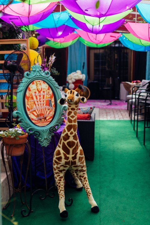Giraffe plush toy in front of colorful decorations and umbrellas hanging from ceiling, with seating area in background.