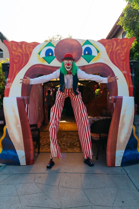 Person dressed as a clown with outstretched arms standing in front of a colorful clown face backdrop.