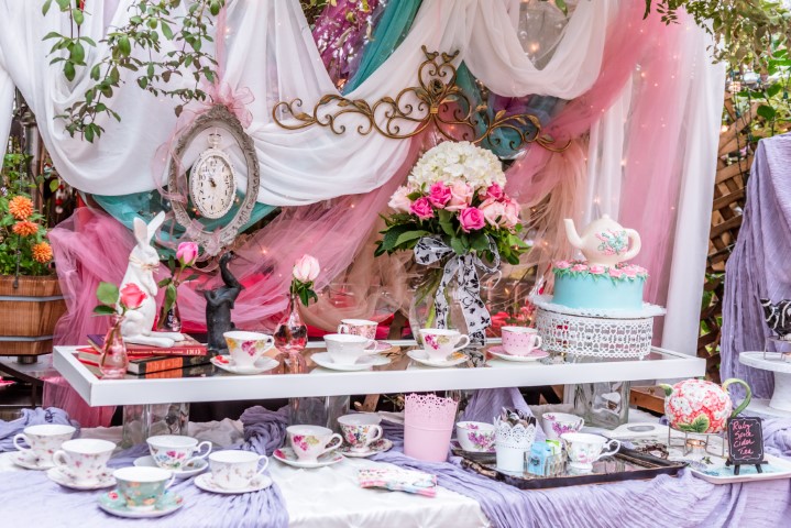 Table with teacups, a cake, flowers, and decorations at a festive event, with colorful drapes and ornaments in background.