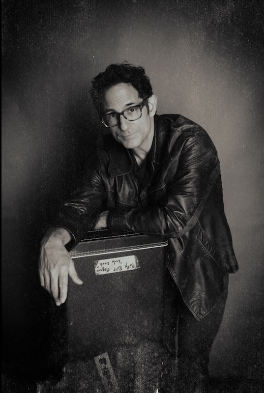 Person with glasses and curly hair leaning on a box, wearing a leather jacket, in black and white.