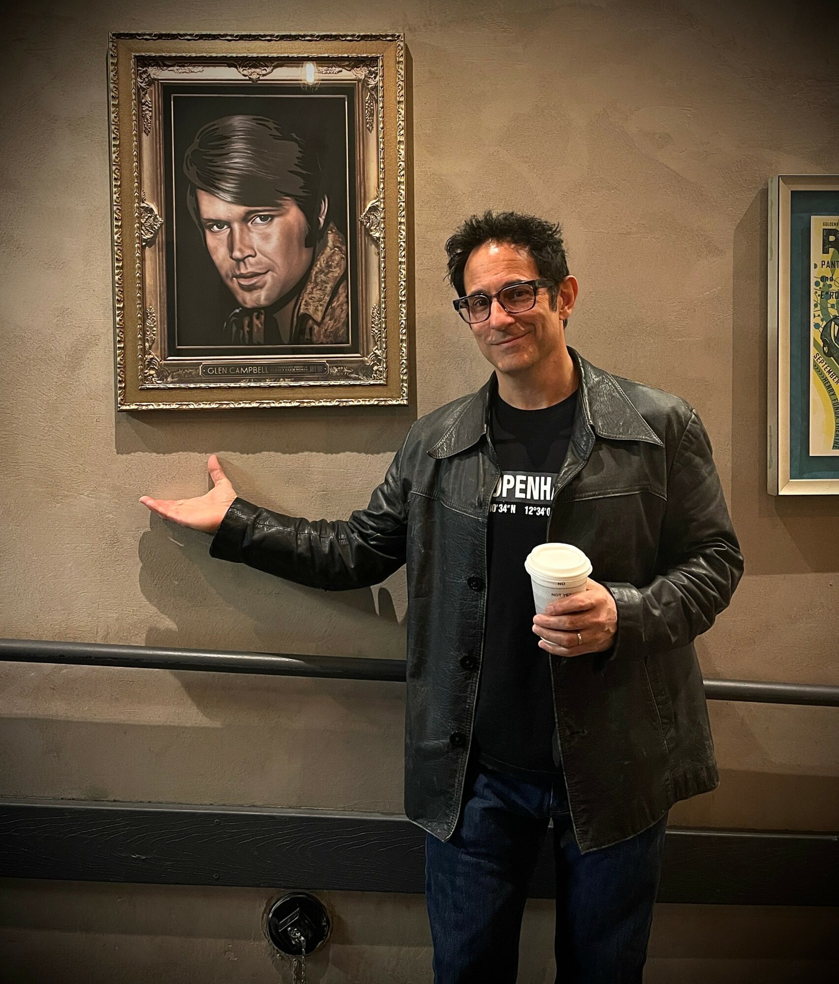 Man with glasses and dark jacket holding a coffee cup, standing in front of a portrait of a man with dark hair.