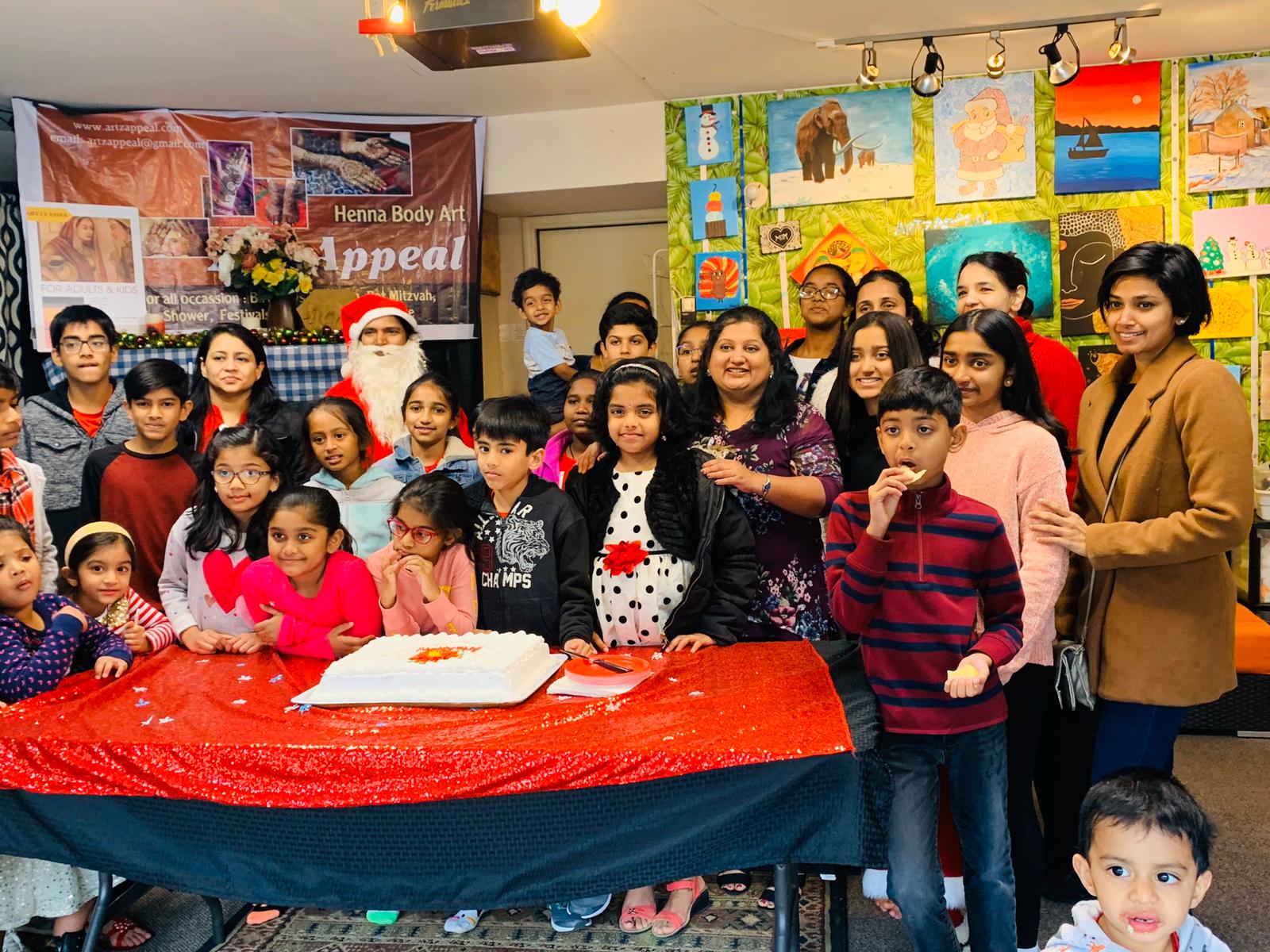 Group of children and adults gathered around a table with a cake, celebrating indoors with colorful artwork on walls.