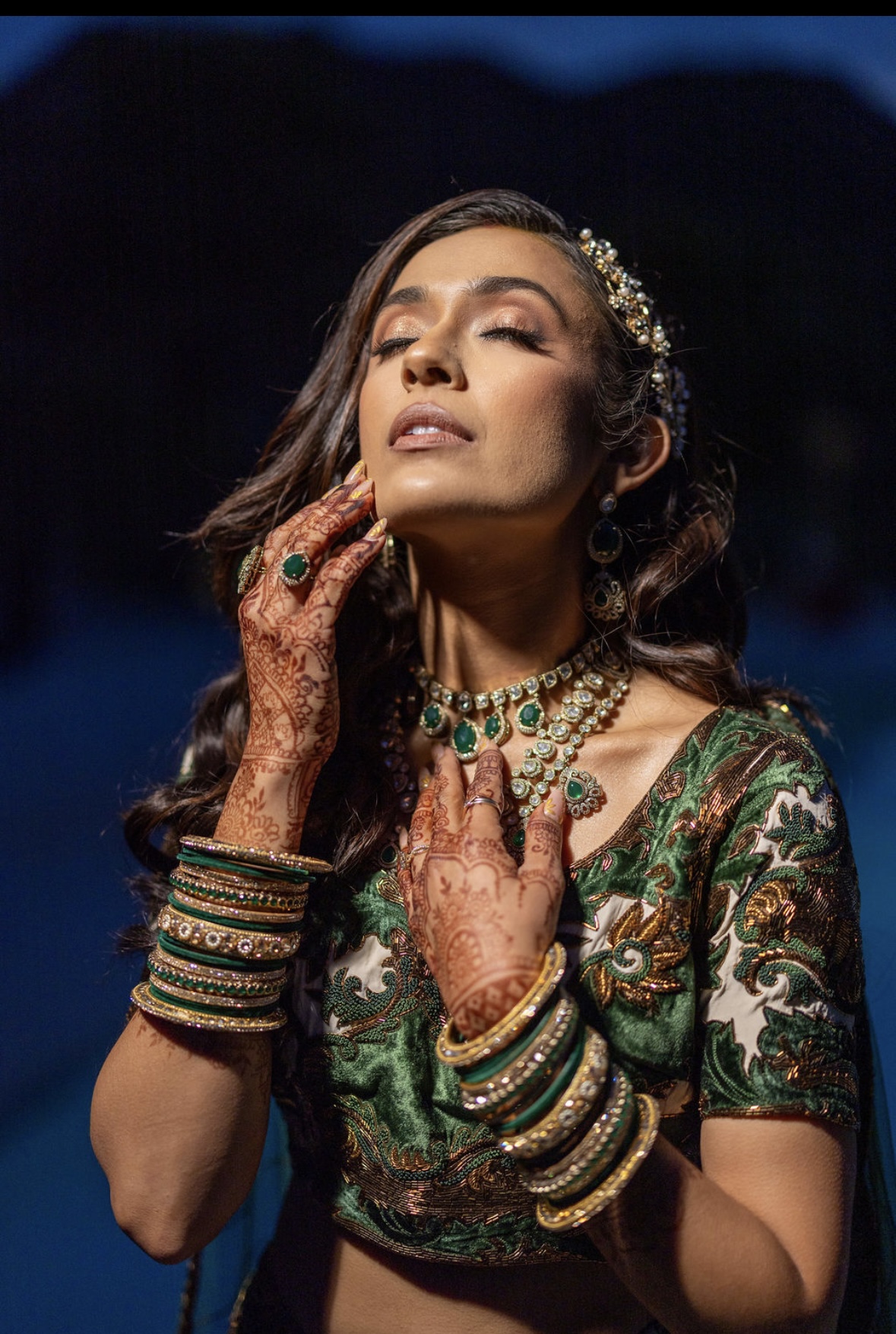 Woman with jewelry and henna on hands, wearing a green patterned dress, posing with eyes closed outdoors at night.